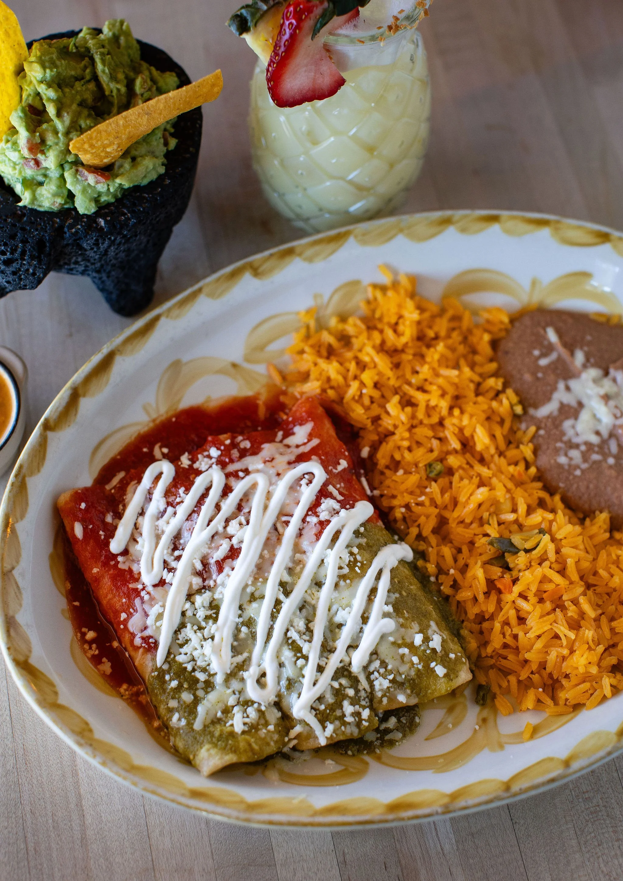 Mexican enchilada plate with red and green enchiladas, Spanish rice, refried beans, guacamole, tortilla chips, and a garnished tropical drink