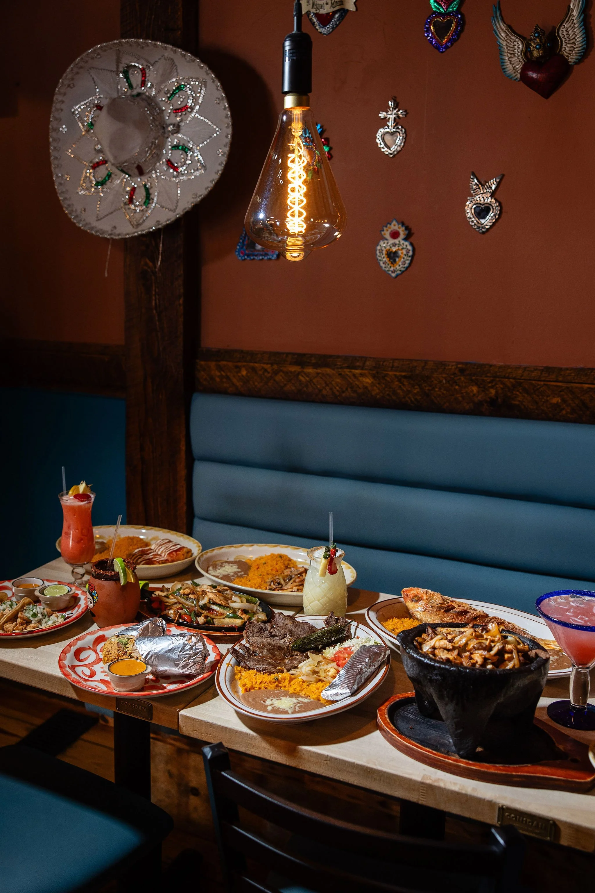 Colorful Mexican restaurant booth with a glowing pendant bulb, heart-shaped wall decorations, blue seating, and a table covered with Mexican dishes, rice, fajitas, grilled meats, and cocktails.