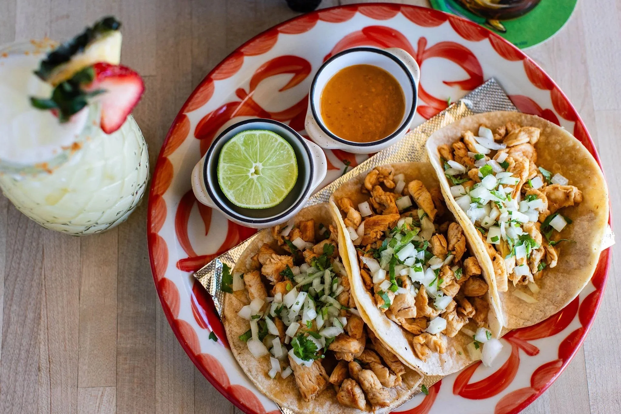 Overhead view of grilled chicken tacos topped with chopped onion and cilantro, served with lime, salsa, and a strawberry-topped drink