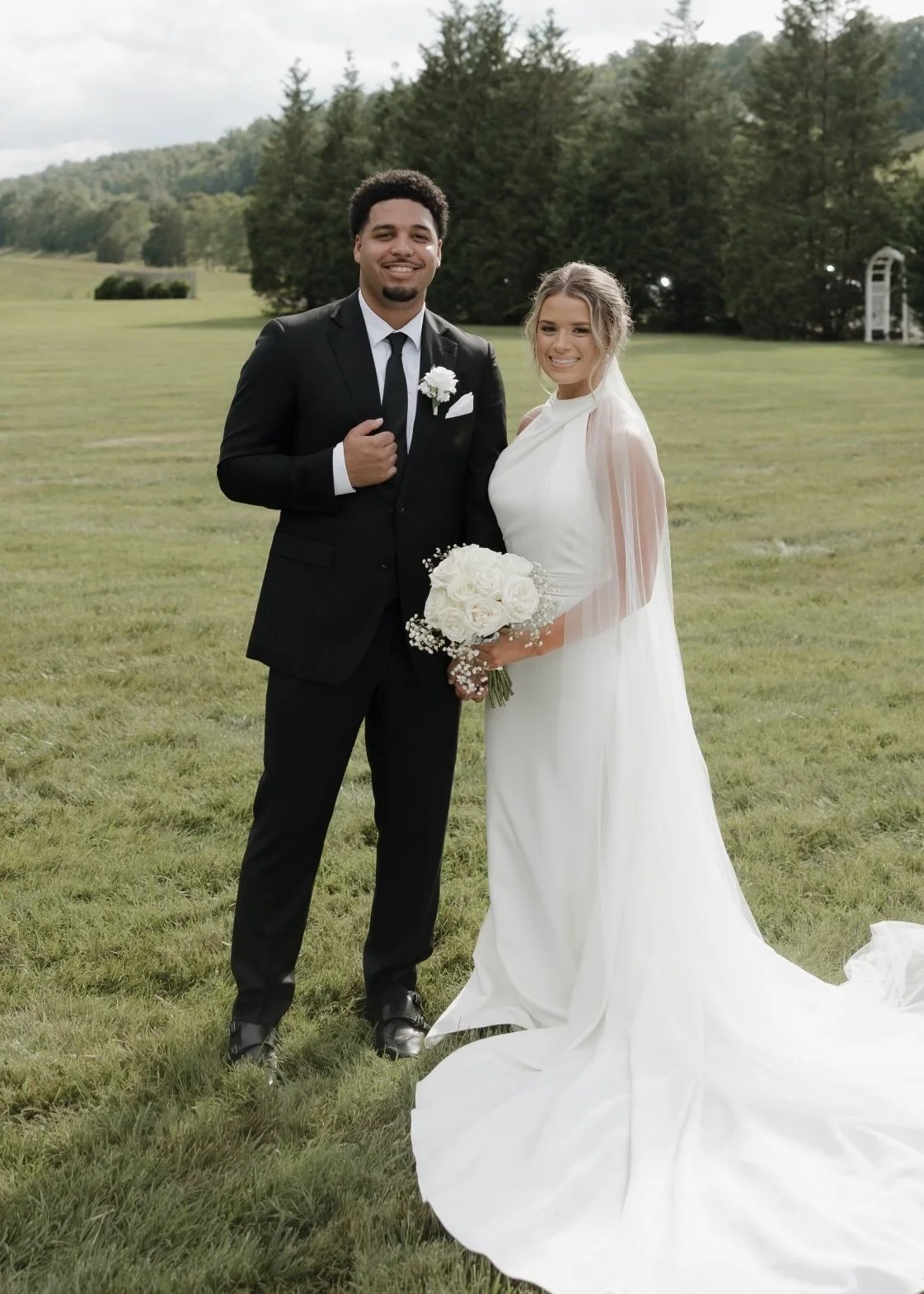 Newlywed couple posing outdoors in a grassy field, with the groom in a black suit and the bride in a white wedding dress holding a bouquet.