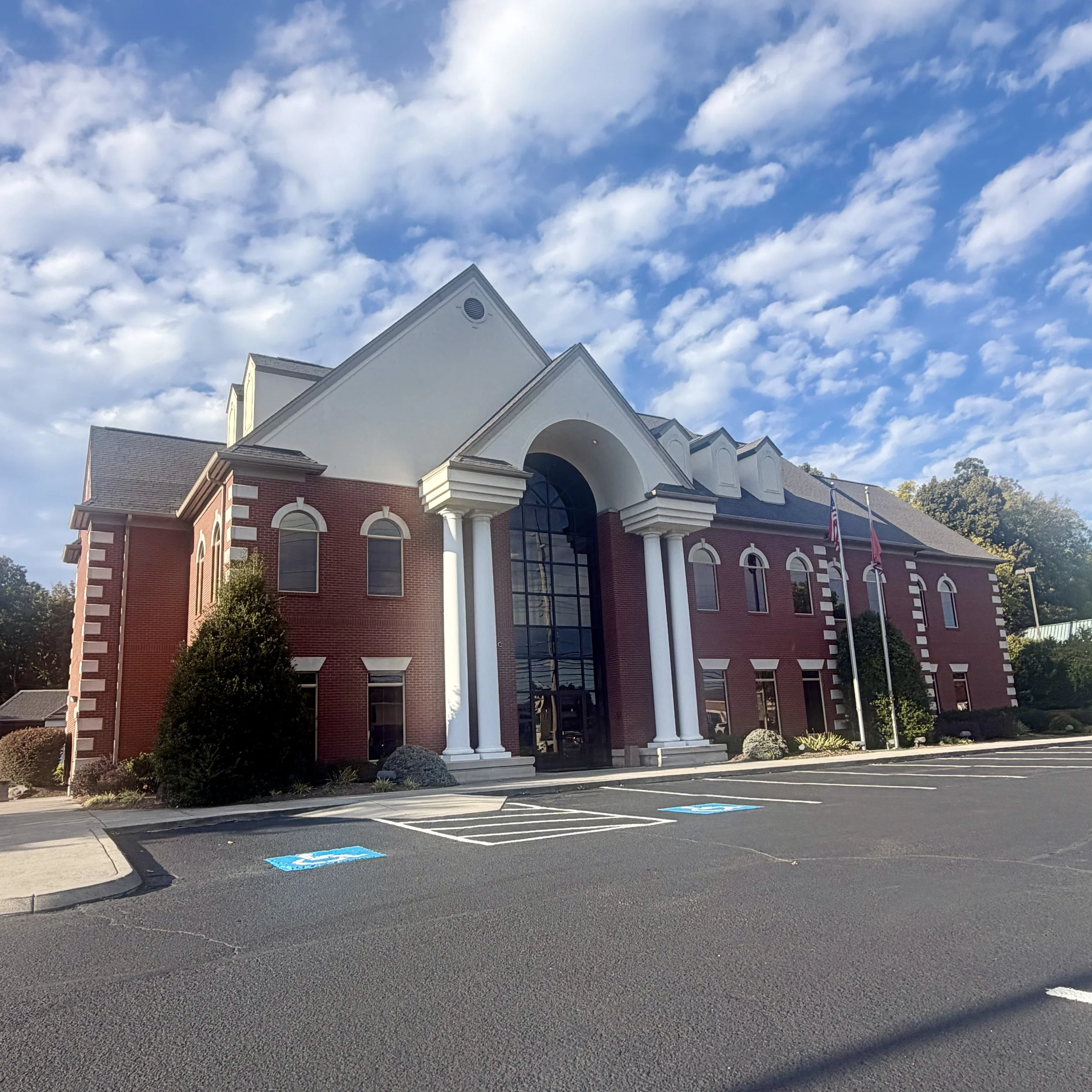 Red-brick building with white columns and American flags beside an accessible parking lot under a partly cloudy blue sky