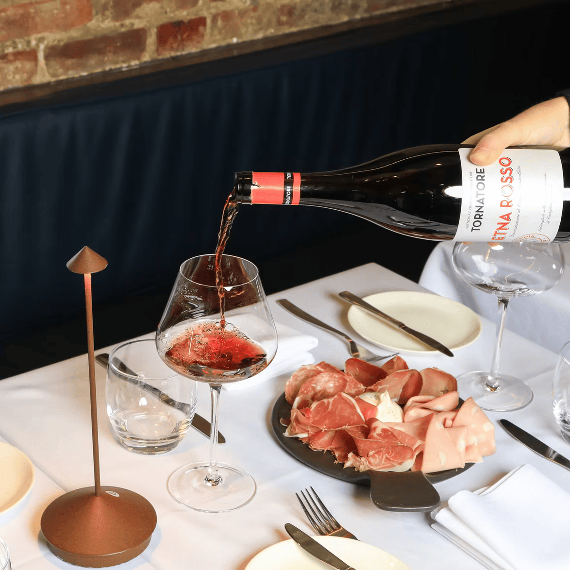 A bottle of Tenuta Tormaresca Negroamaro rosé wine is being poured into a wine glass at a restaurant table with cured meats.