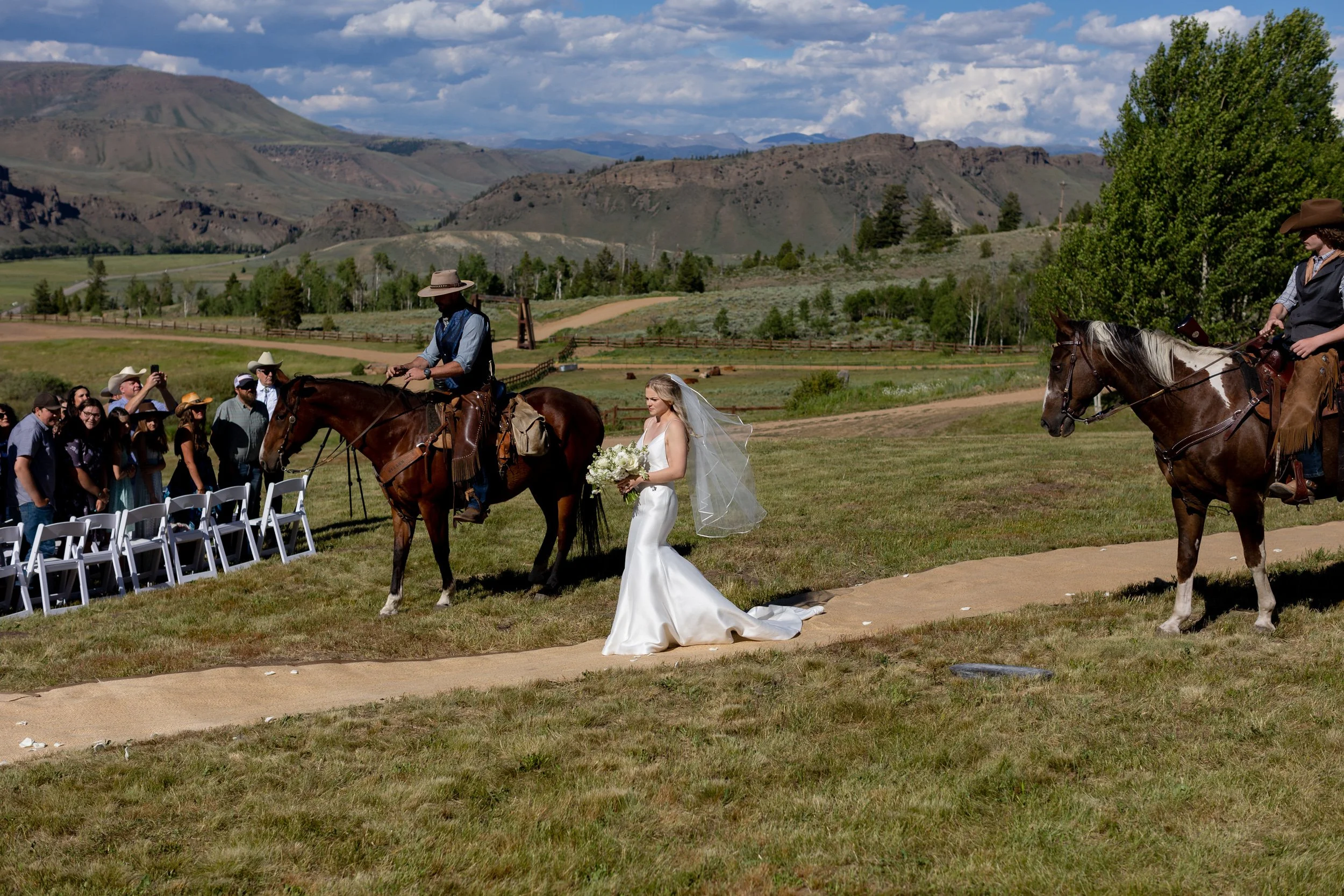 Bride in a white gown walking a dirt path between riders on horseback in a meadow in Hot Sulphur Springs, Colorado
