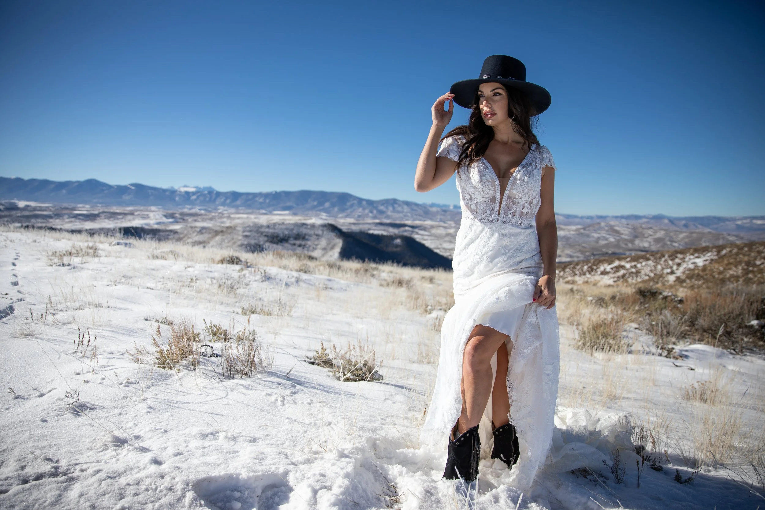 Woman in a black hat and white gown posing on snowy ground with mountains in the distance in Hot Sulphur Springs, Colorado