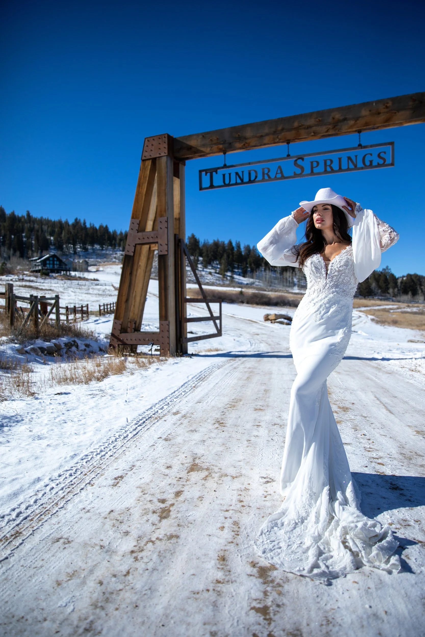 Bride in a white gown and hat on a snowy road near a timber entrance arch at Tundra Springs, Colorado mountain wedding venue