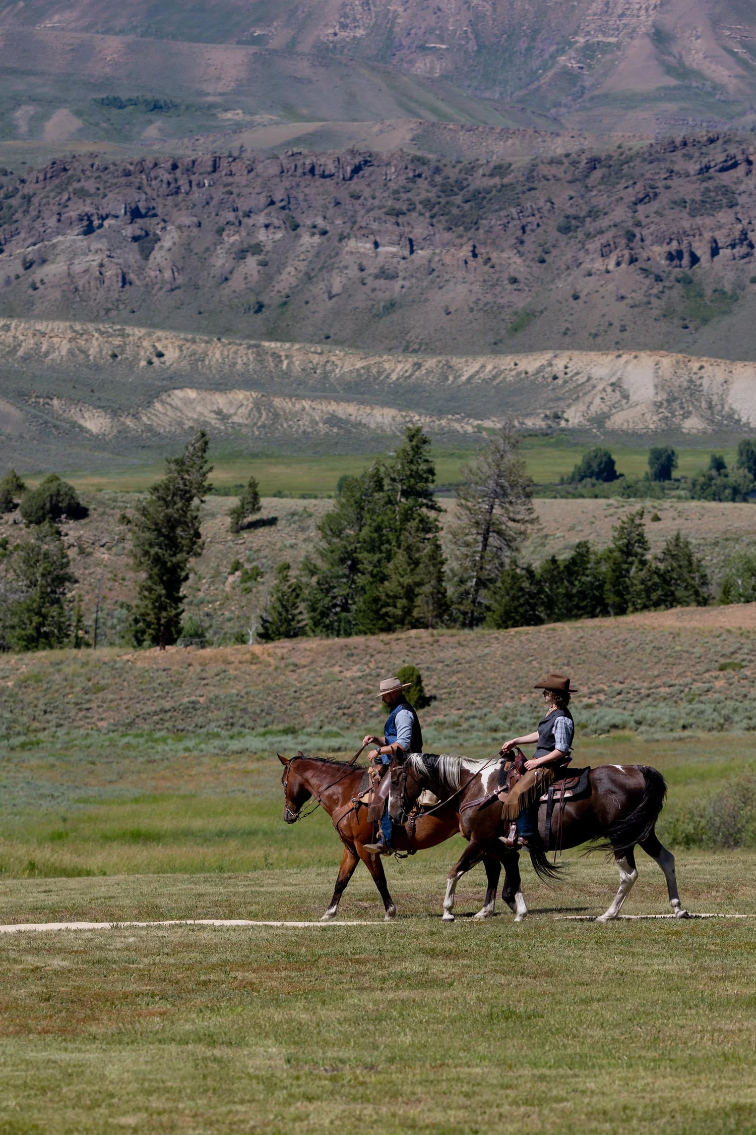 Two riders on horseback crossing a grassy meadow with rugged hills behind at Tundra Springs, Colorado mountain wedding venue