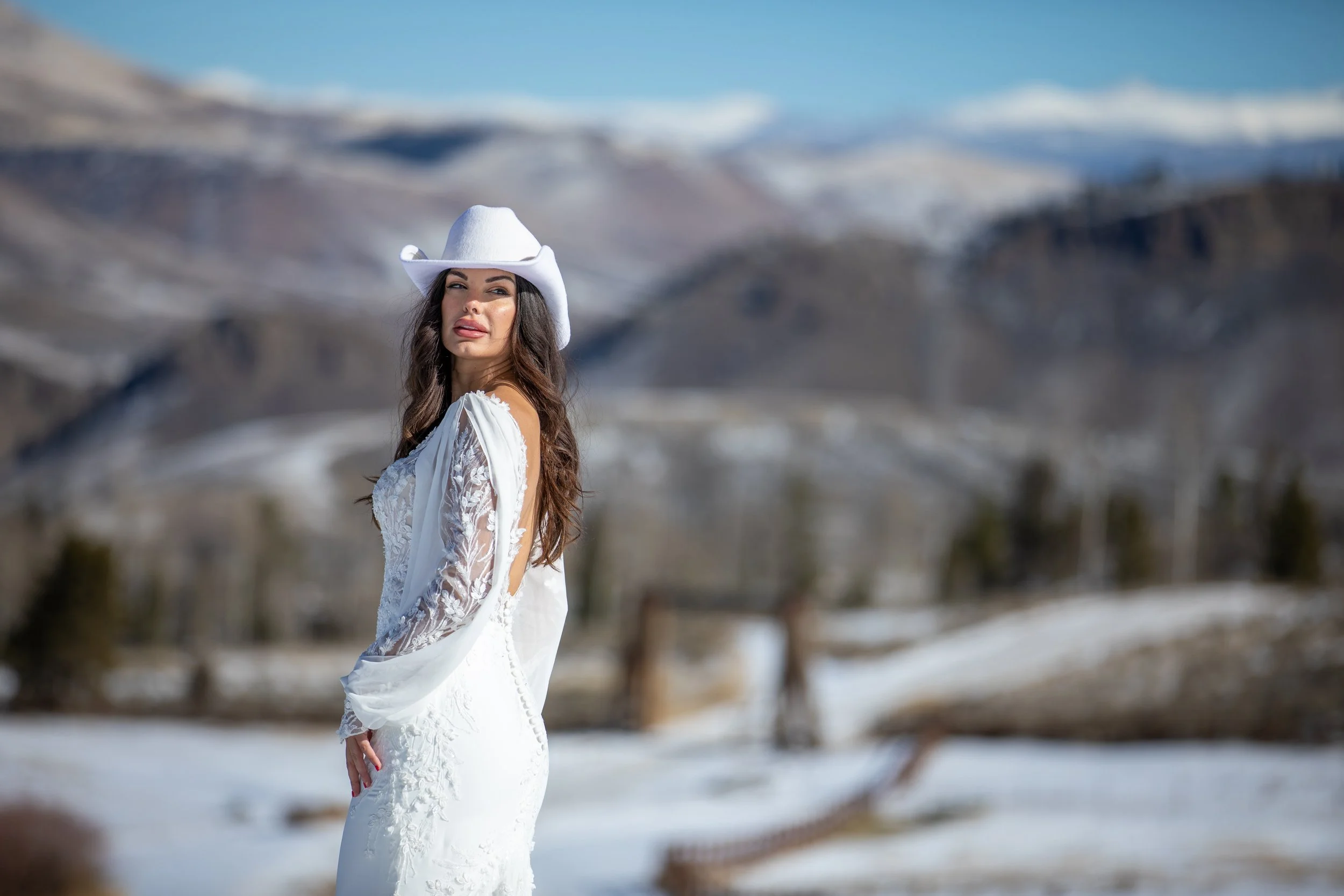 Bride in a white hat and long-sleeved lace gown in a snowy valley with mountain ridges in Hot Sulphur Springs, Colorado