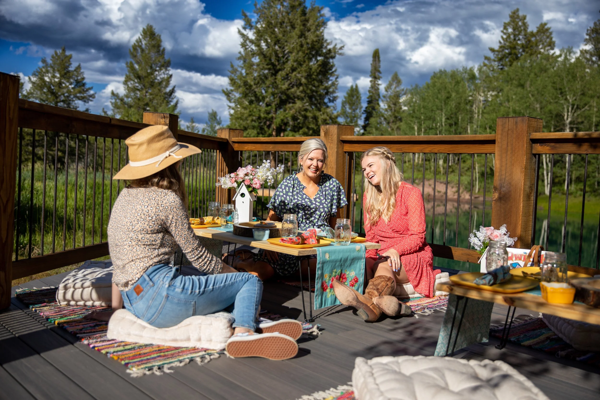 Friends relaxing around a wooden table on a mountain deck with pine trees and blue sky in Hot Sulphur Springs, Colorado