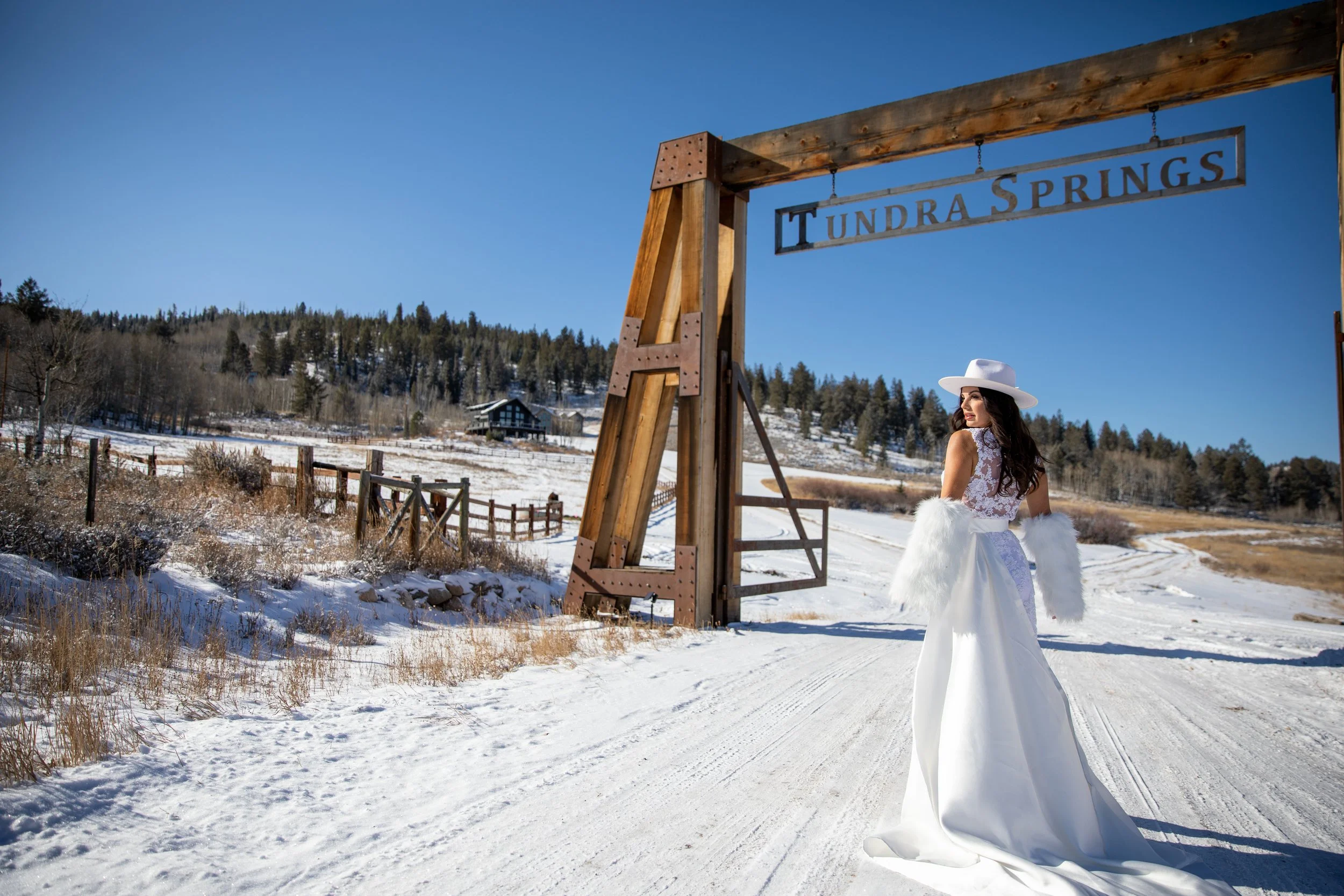 Bride in a white fur wrap beside the Tundra Springs gate sign on a snowy road in Hot Sulphur Springs, Colorado