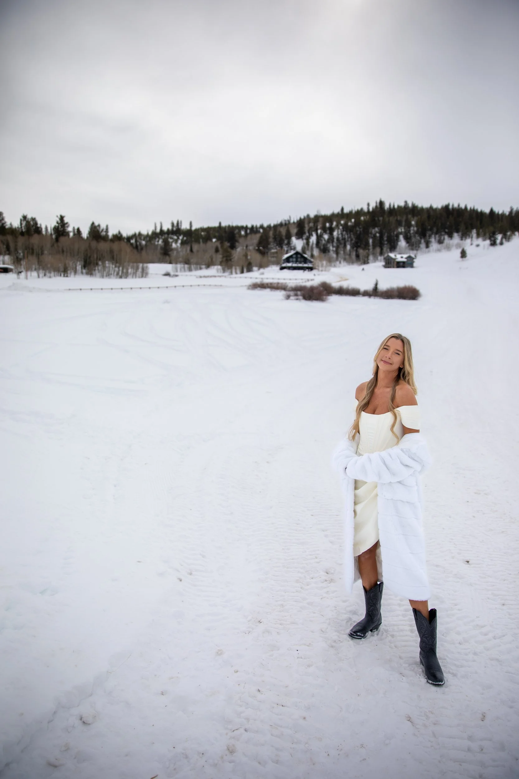 Woman in a long white coat standing in a wide snowfield below the treeline at Tundra Springs, Colorado mountain wedding venue