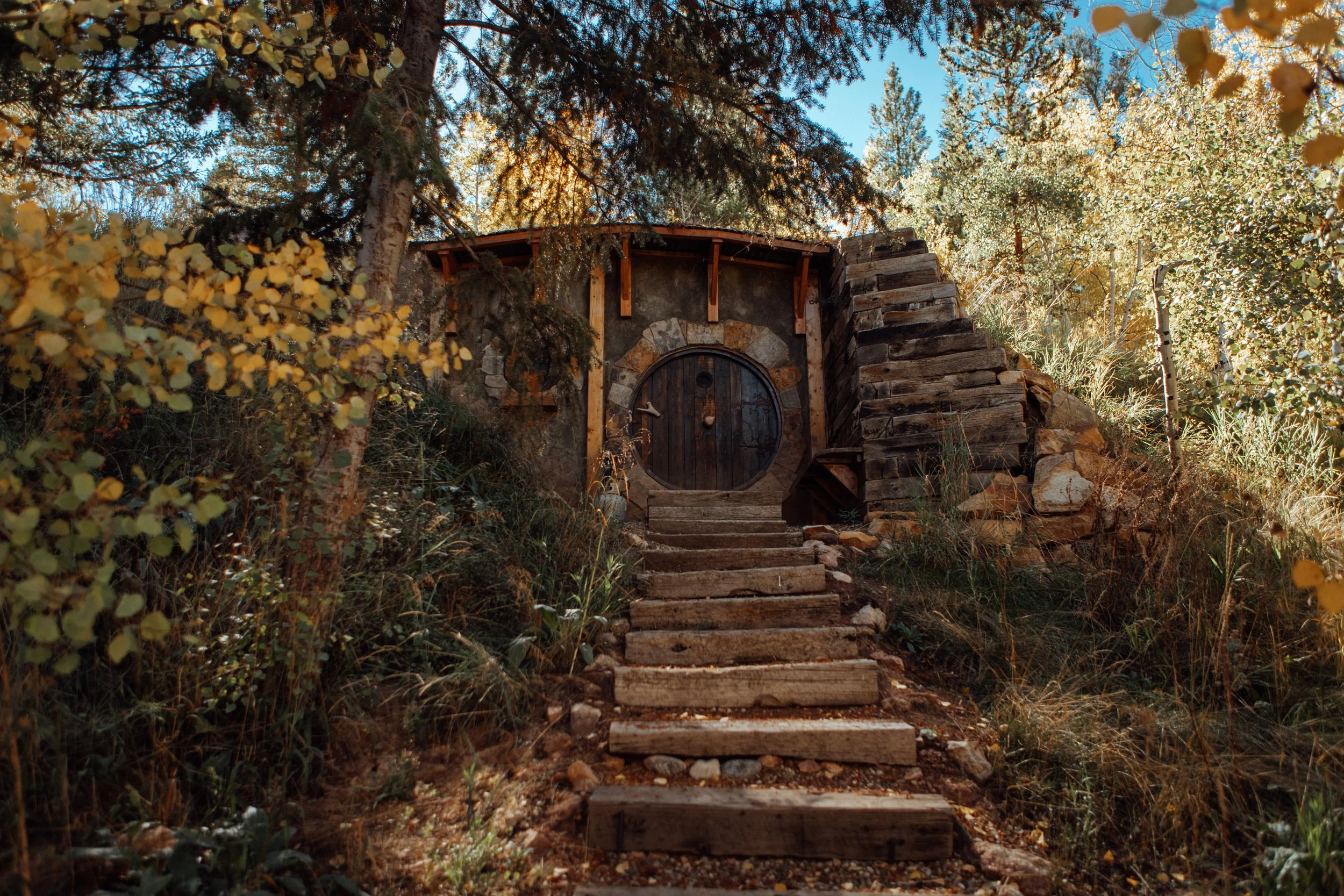 Round wooden Hobbit Hole door atop stone steps amid autumn foliage at Tundra Springs, Colorado mountain wedding venue
