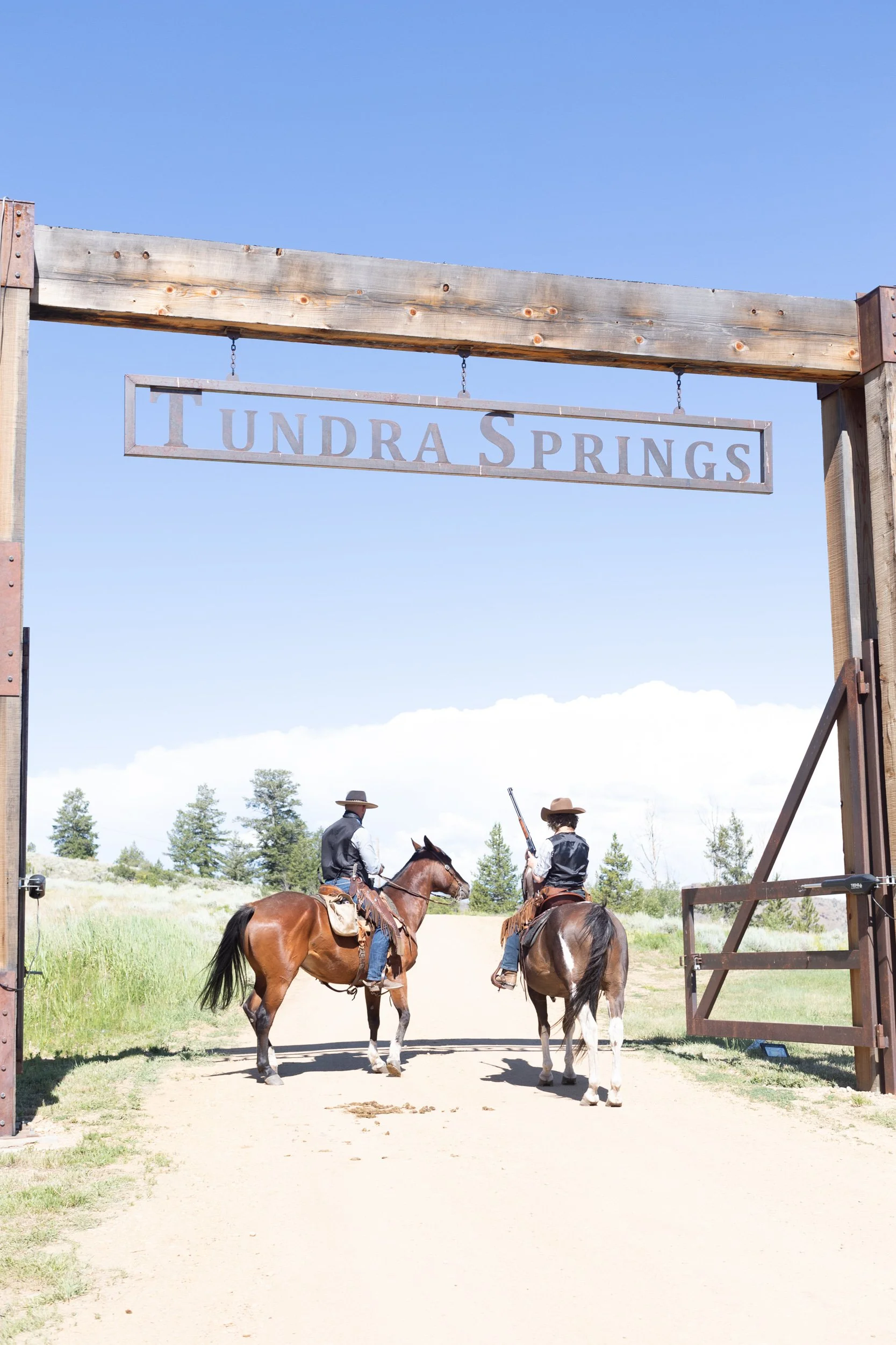 Two riders on horseback passing under the Tundra Springs entrance gate on a dirt road in Hot Sulphur Springs, Colorado