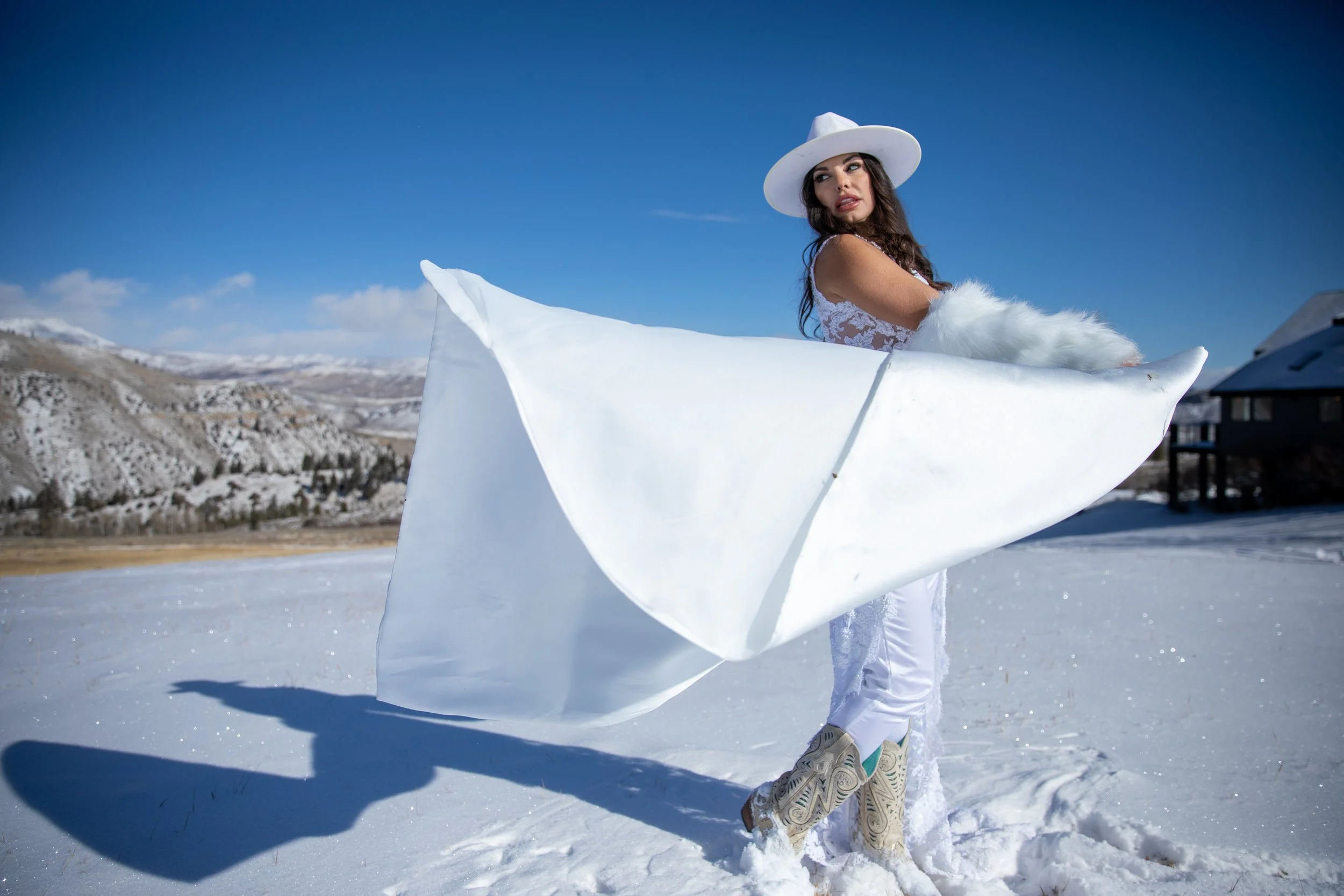 Bride in a white hat and gown holding a billowing white wrap on a snowy field in Hot Sulphur Springs, Colorado