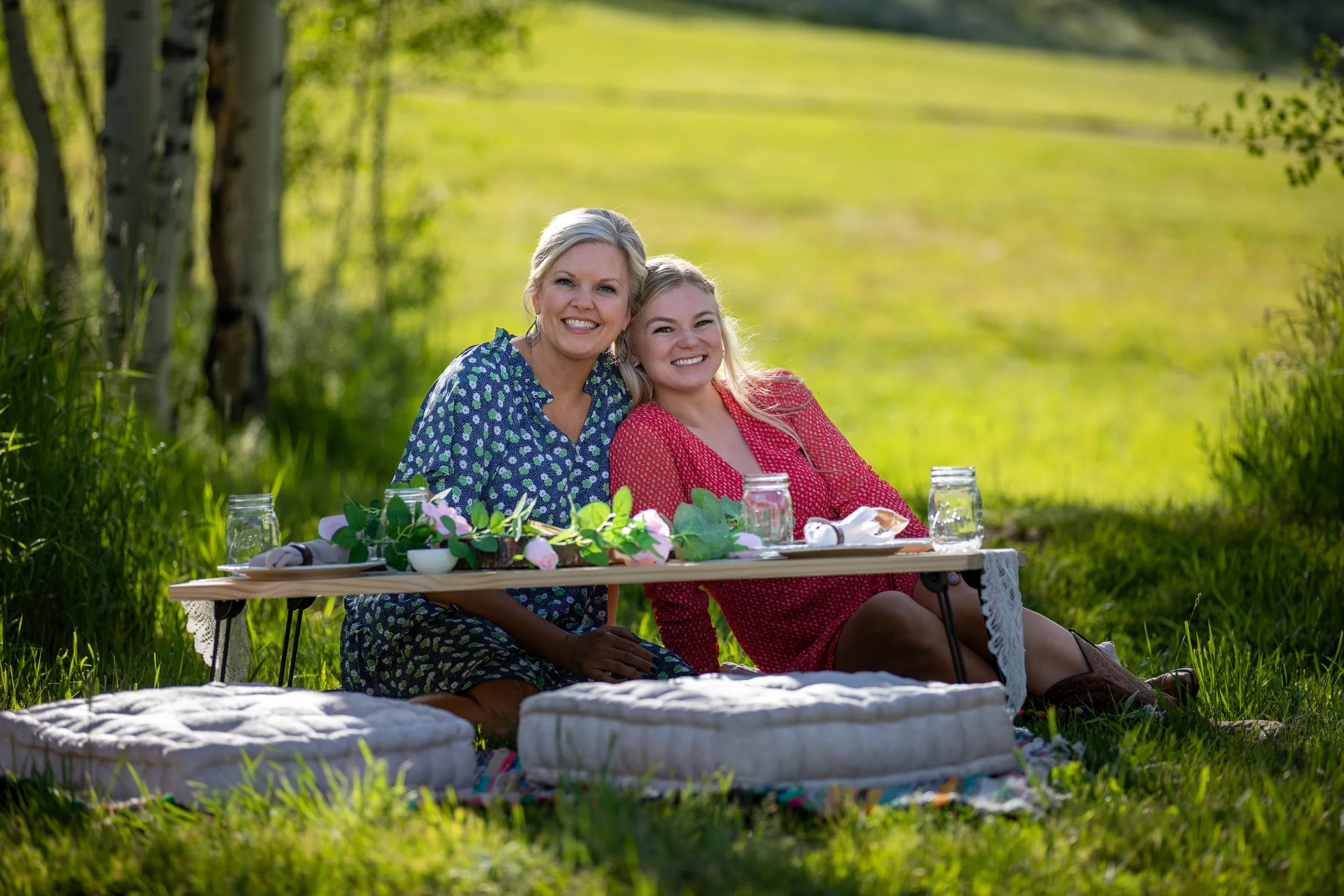 Two women seated on cushions at a styled low table on a sunny green lawn at Tundra Springs, Colorado mountain wedding venue