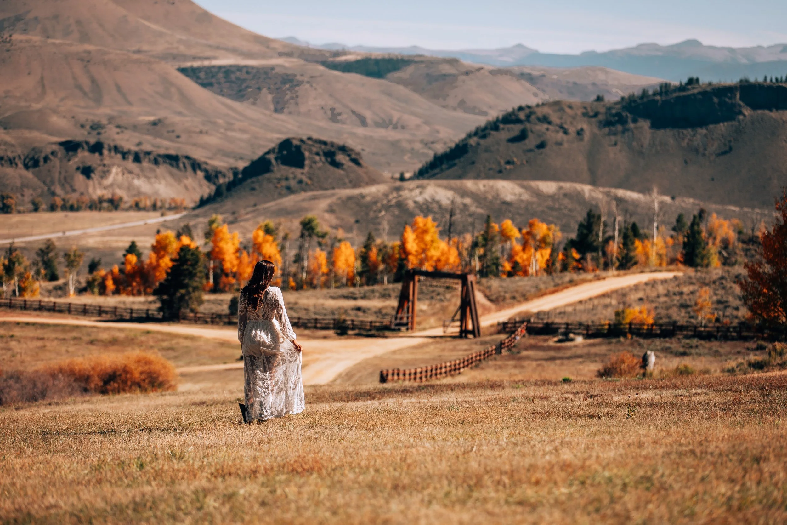 Woman in a long dress walking a dirt road past a wooden bridge in autumn at Tundra Springs, Colorado mountain wedding venue