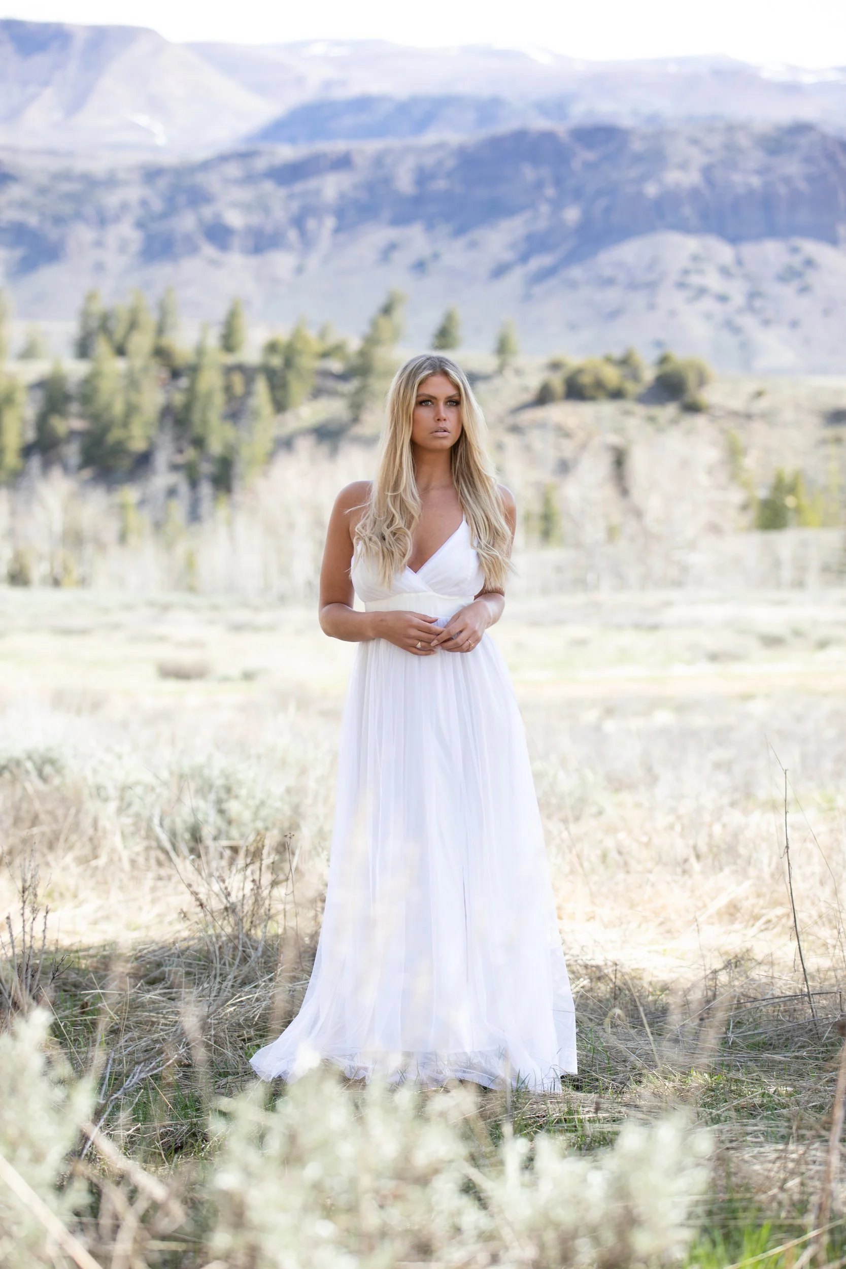 Bride in a flowing white lace gown standing in tall meadow grass near pine trees in Hot Sulphur Springs, Colorado