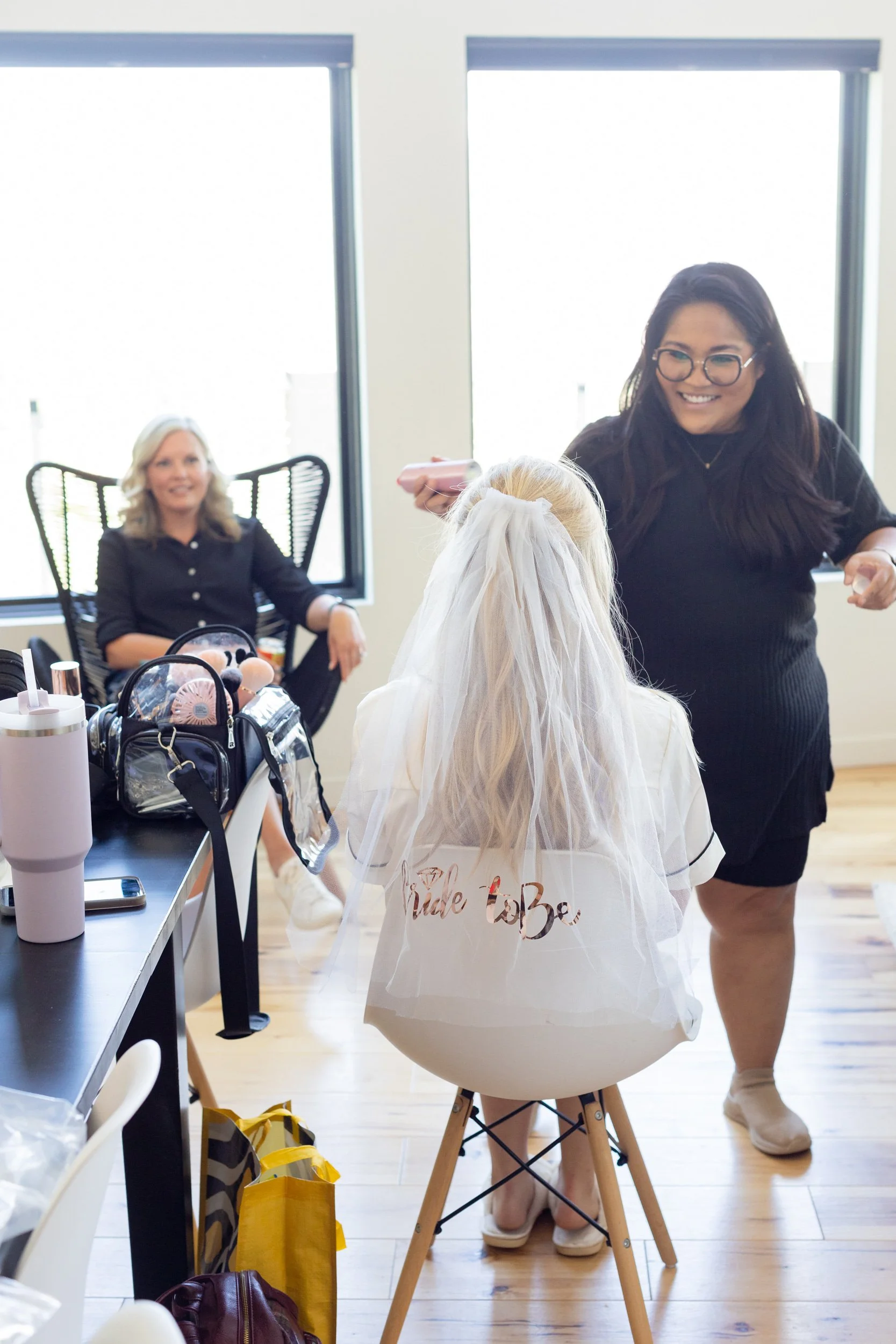 Bride in a white robe seated in a bright room while a friend in black walks past in Hot Sulphur Springs, Colorado