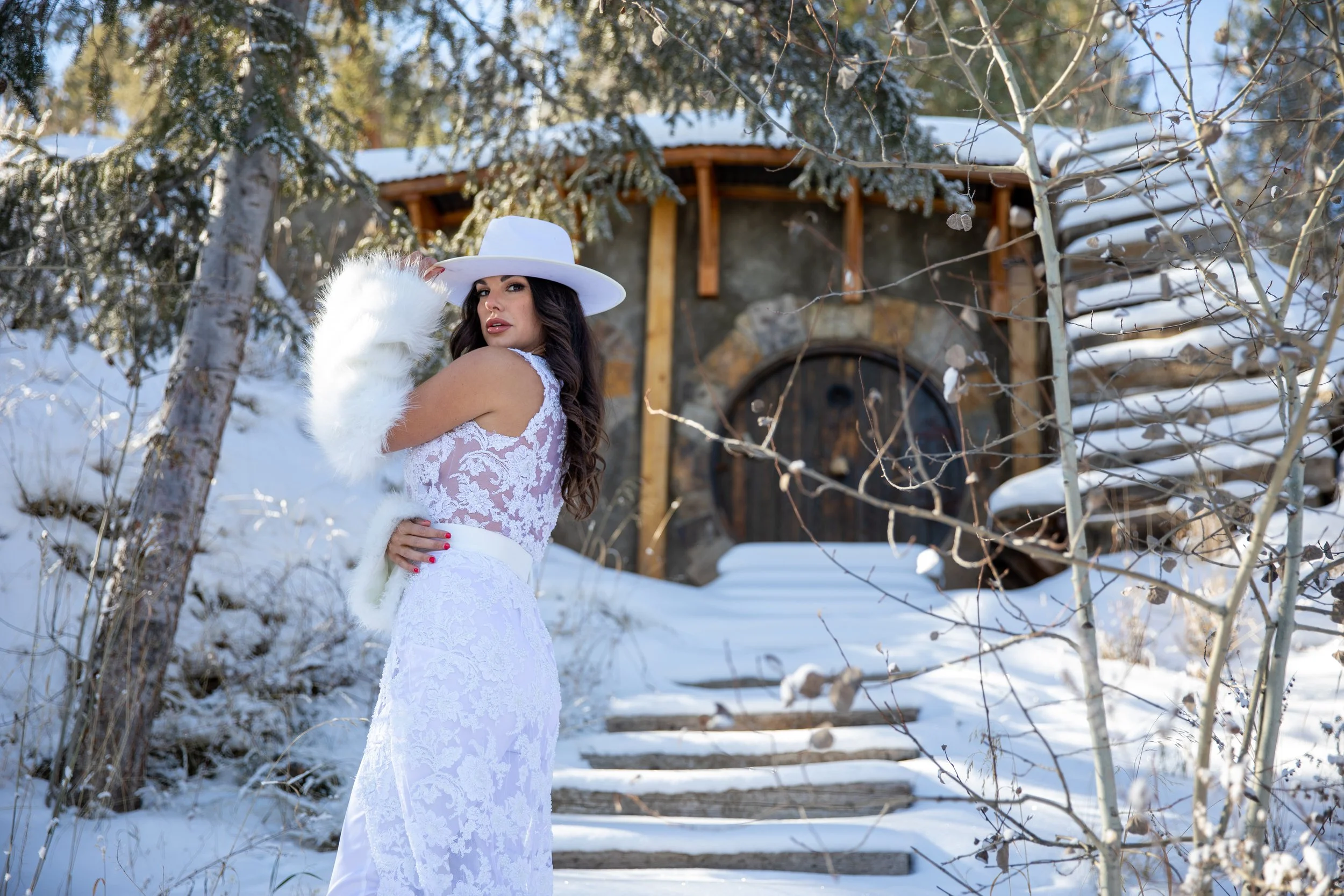 Bride in a fur stole and hat on stone steps by a round stone doorway in a snowy grove in Hot Sulphur Springs, Colorado