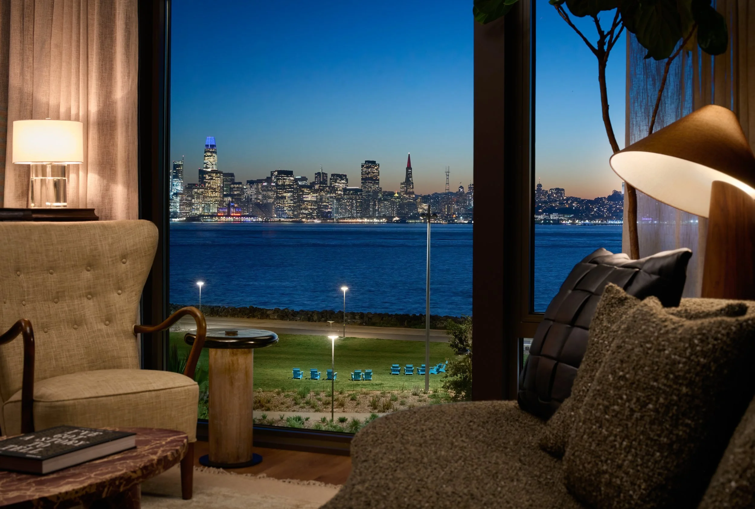 Lounge armchair beside a window framing the lit San Francisco skyline at dusk, 490 Avenue of the Palms, Treasure Island