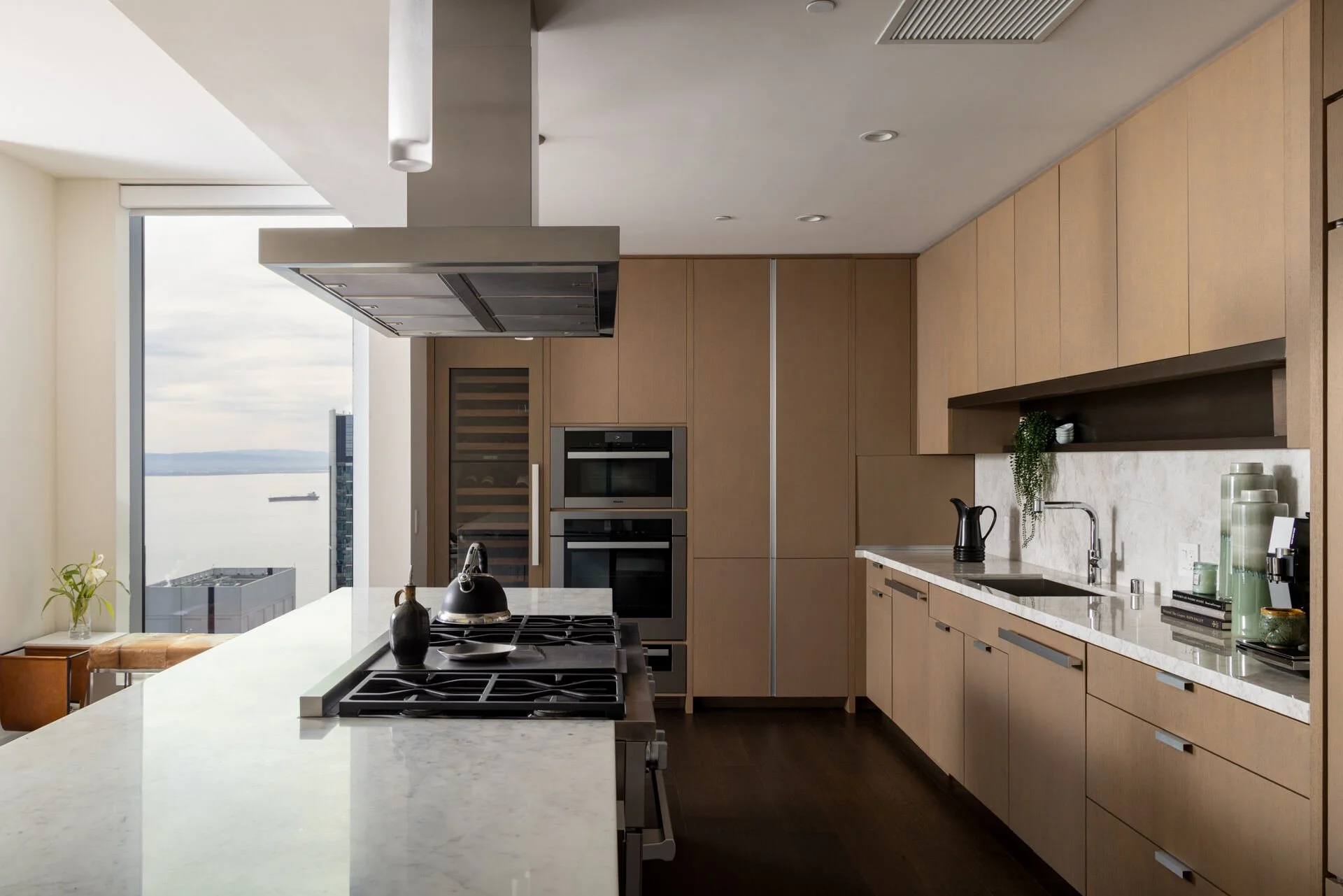 Modern luxury kitchen with wood cabinets, marble island, gas range, wine refrigerator, and waterfront view