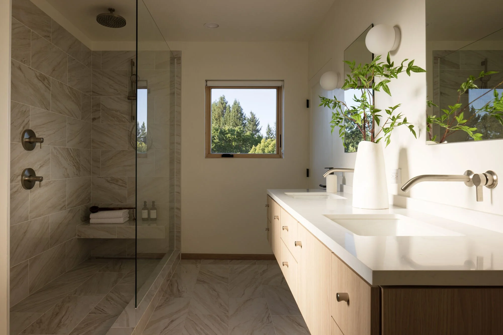 Modern bathroom with marble tile walk-in shower, double vanity, large mirror, potted plant, and tree-view window