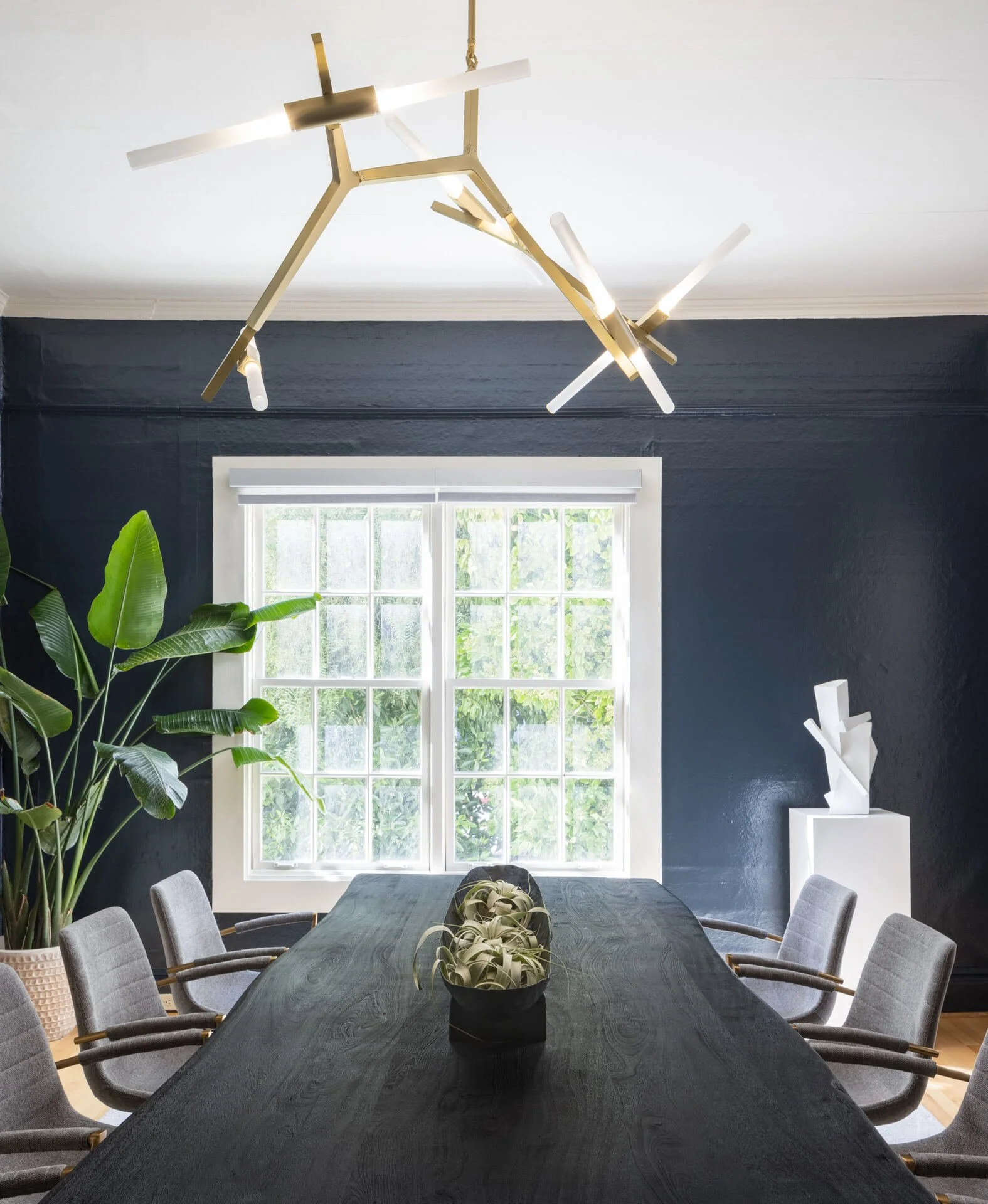 Modern dining room with a large black table, gray upholstered chairs, geometric pendant light, dark walls, green plant, and bright window