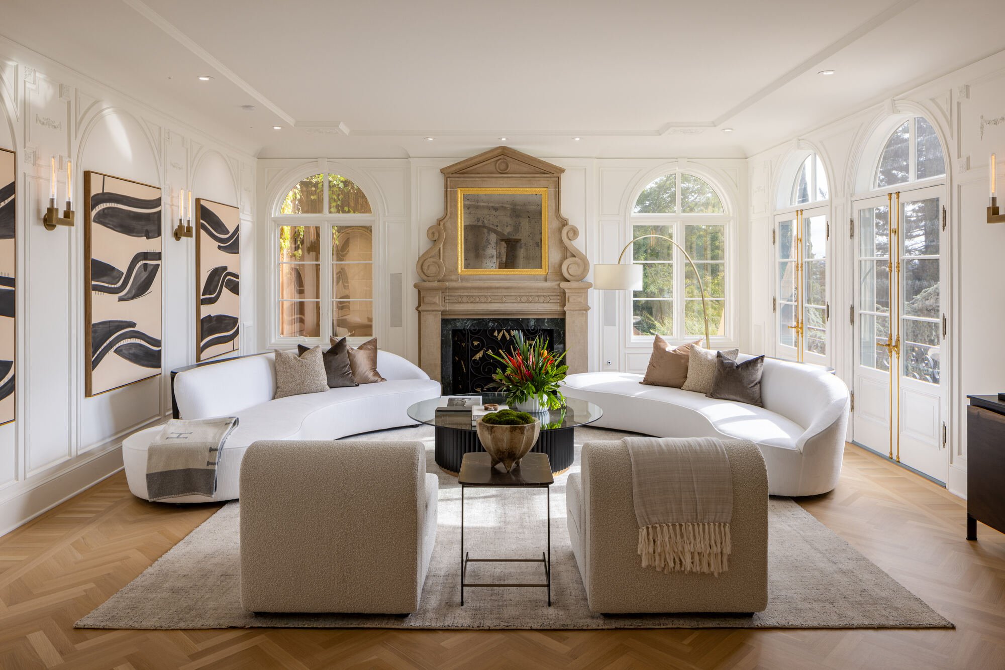Elegant white living room with curved sofas, stone fireplace, arched windows, abstract artwork, and glass coffee table