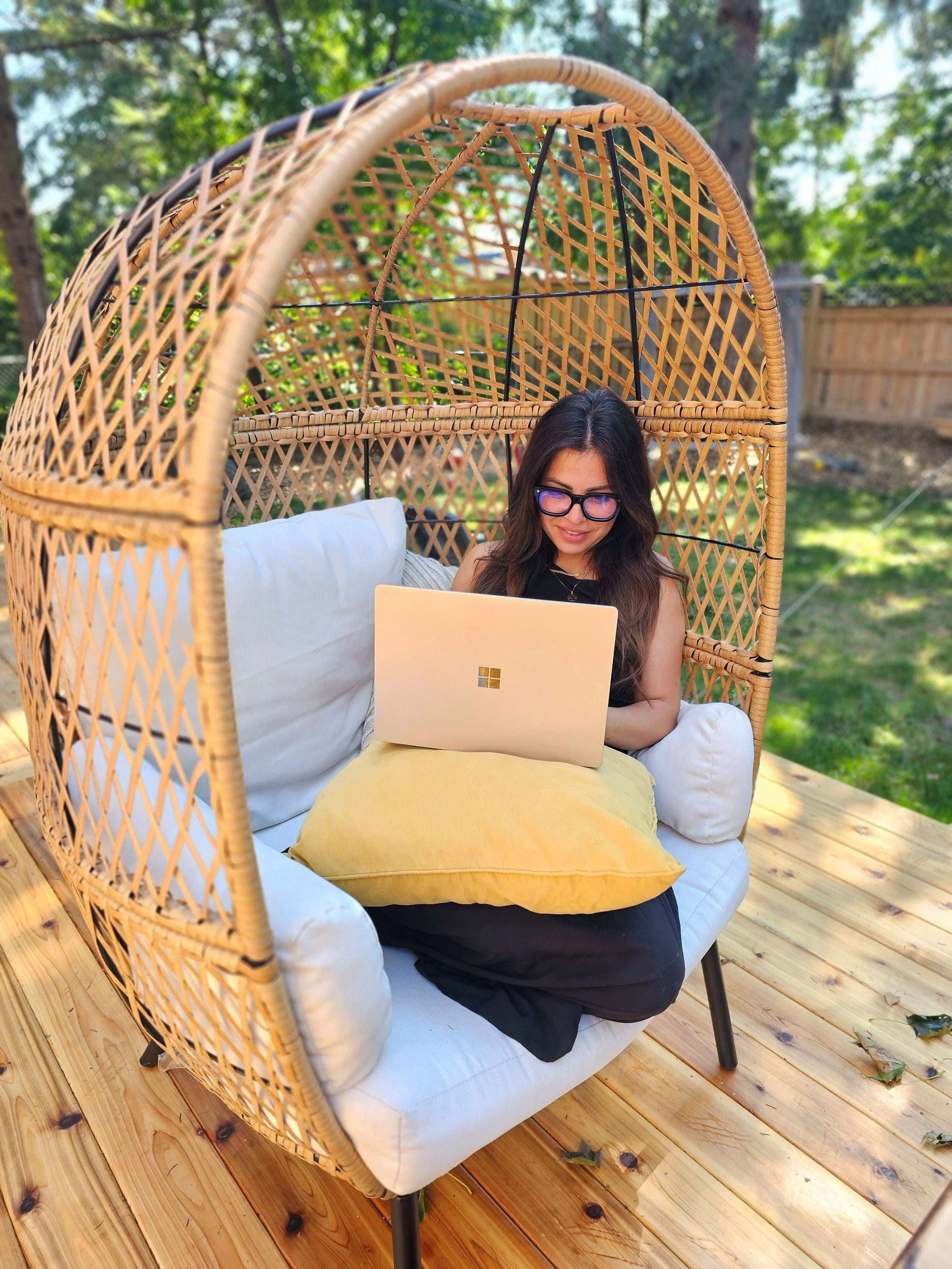 Woman wearing glasses works on a laptop in a woven hanging chair on a sunny wooden deck.