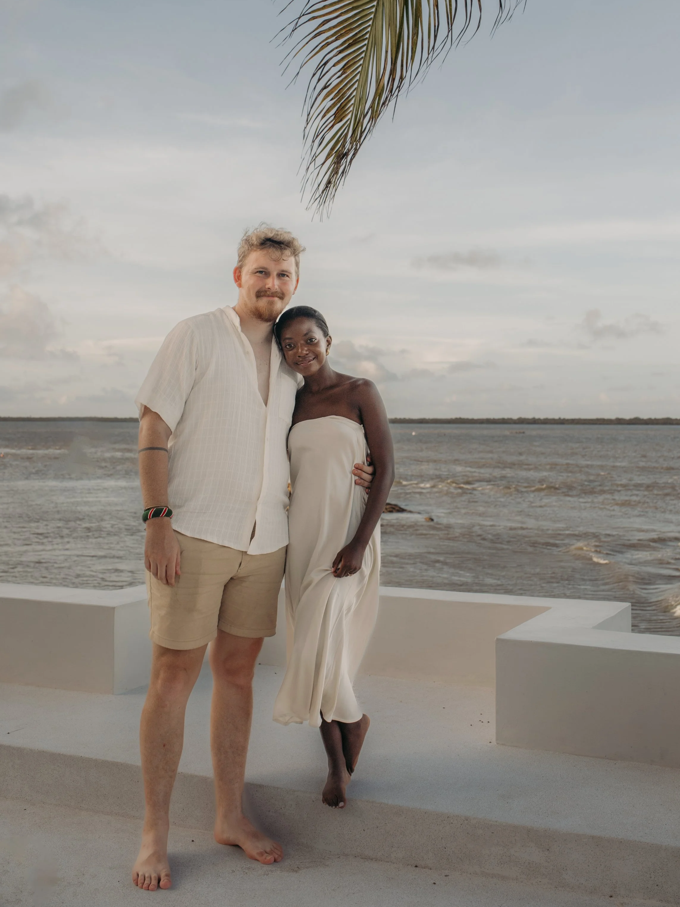 Couple in white clothing embracing beside the ocean beneath a palm tree