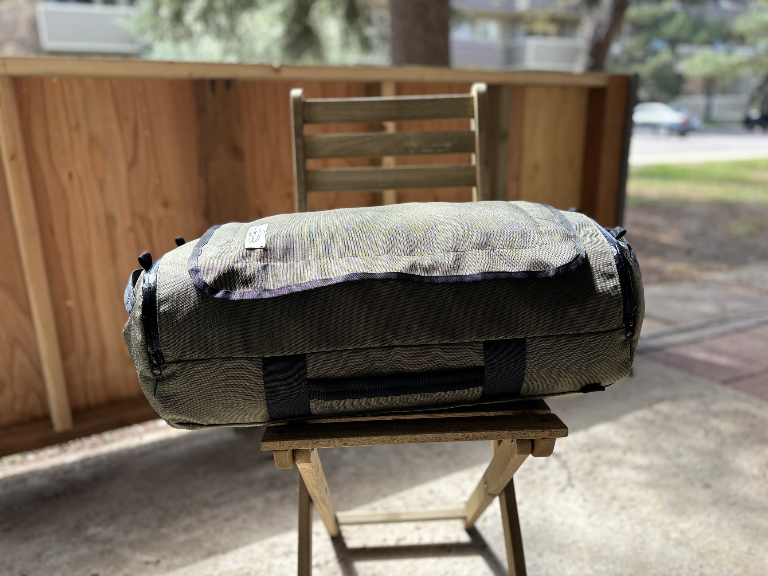 Large olive green duffel bag with black straps displayed on a wooden folding stool outdoors