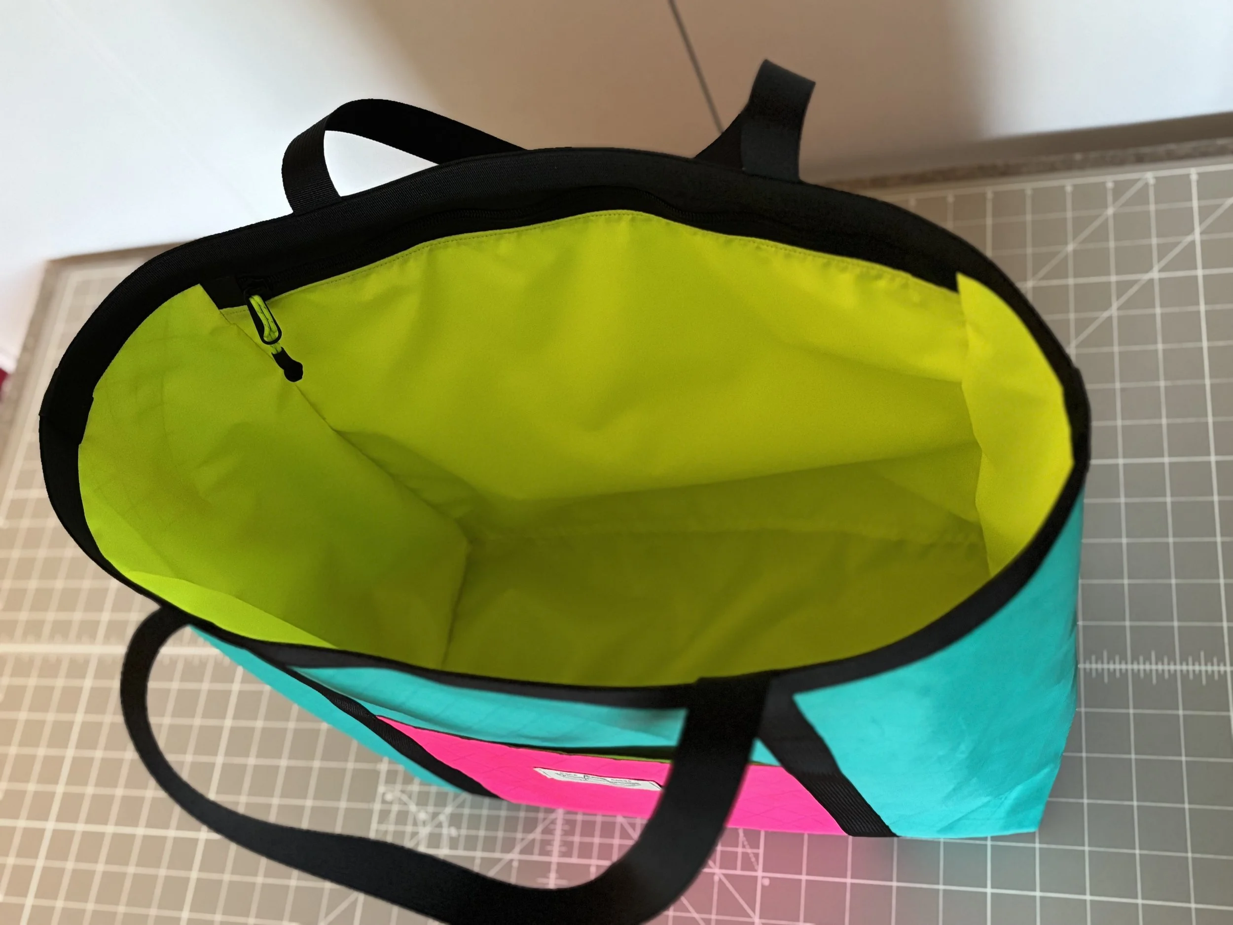 Open handmade color-blocked tote bag with neon green lining, turquoise and pink exterior, and black straps on a cutting mat