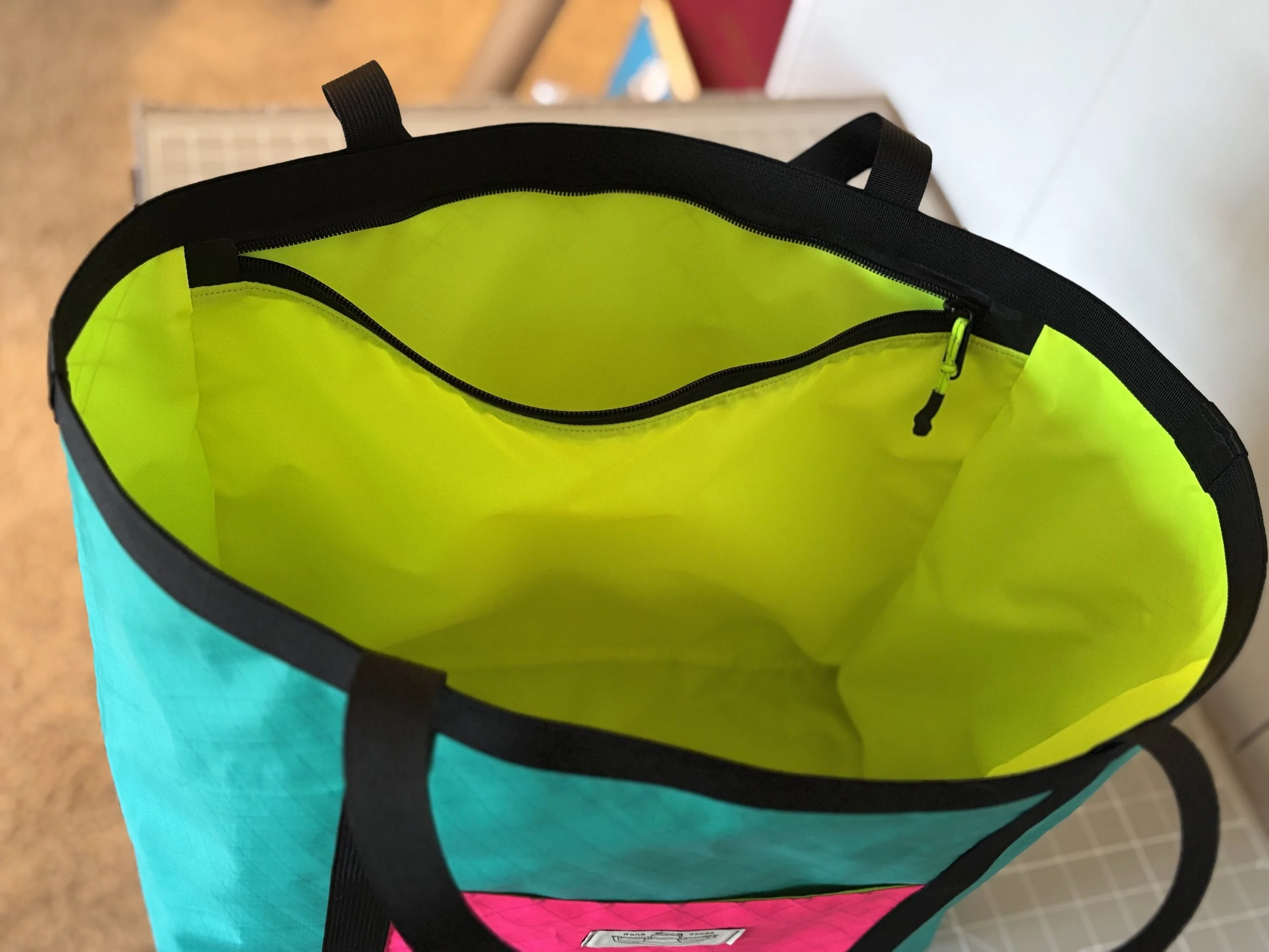 Open turquoise tote bag with a bright lime-green interior, black handles, and zippered pocket.