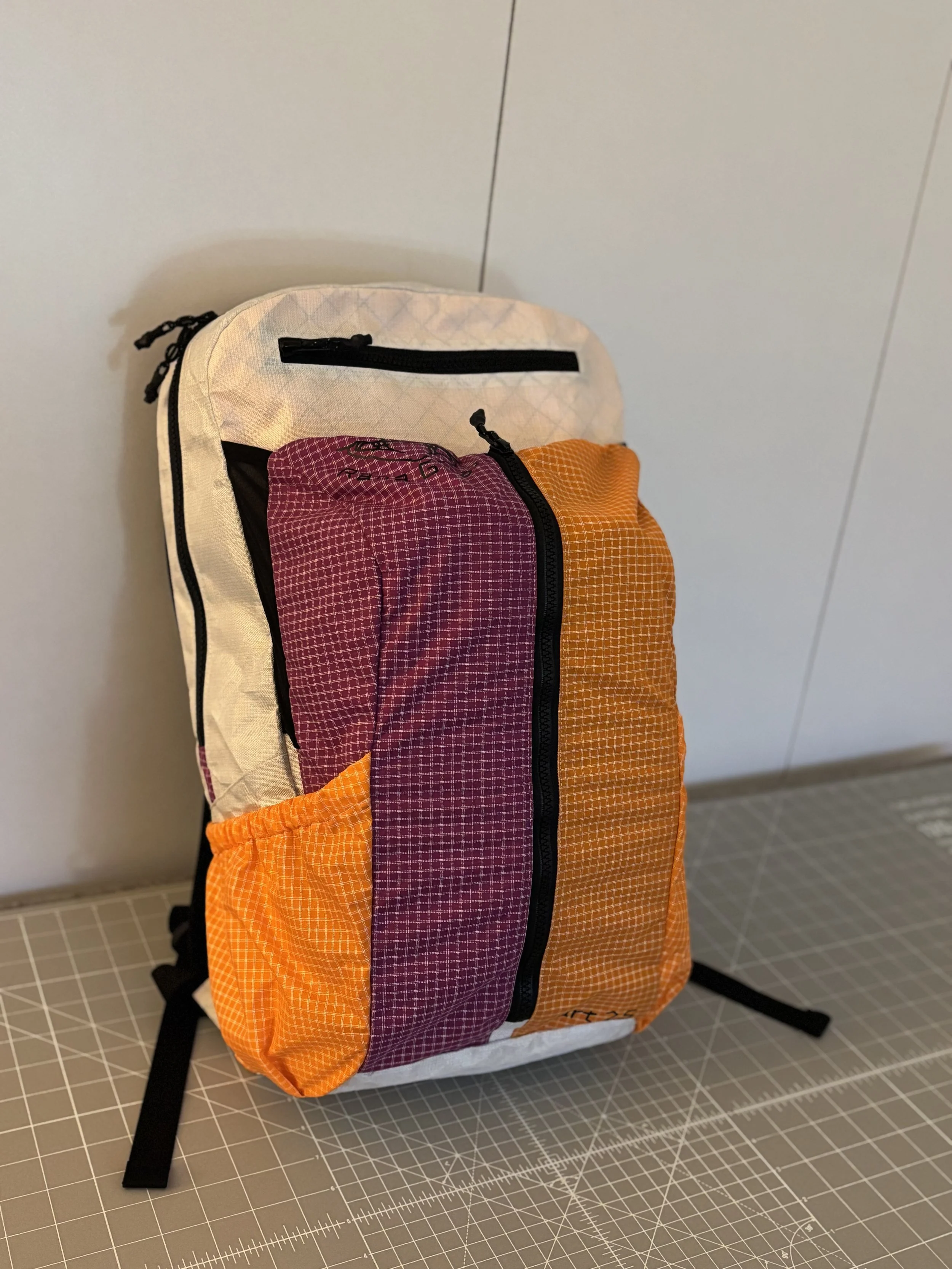 Handmade orange and purple plaid backpack with a cream lining and black zippers on a cutting mat