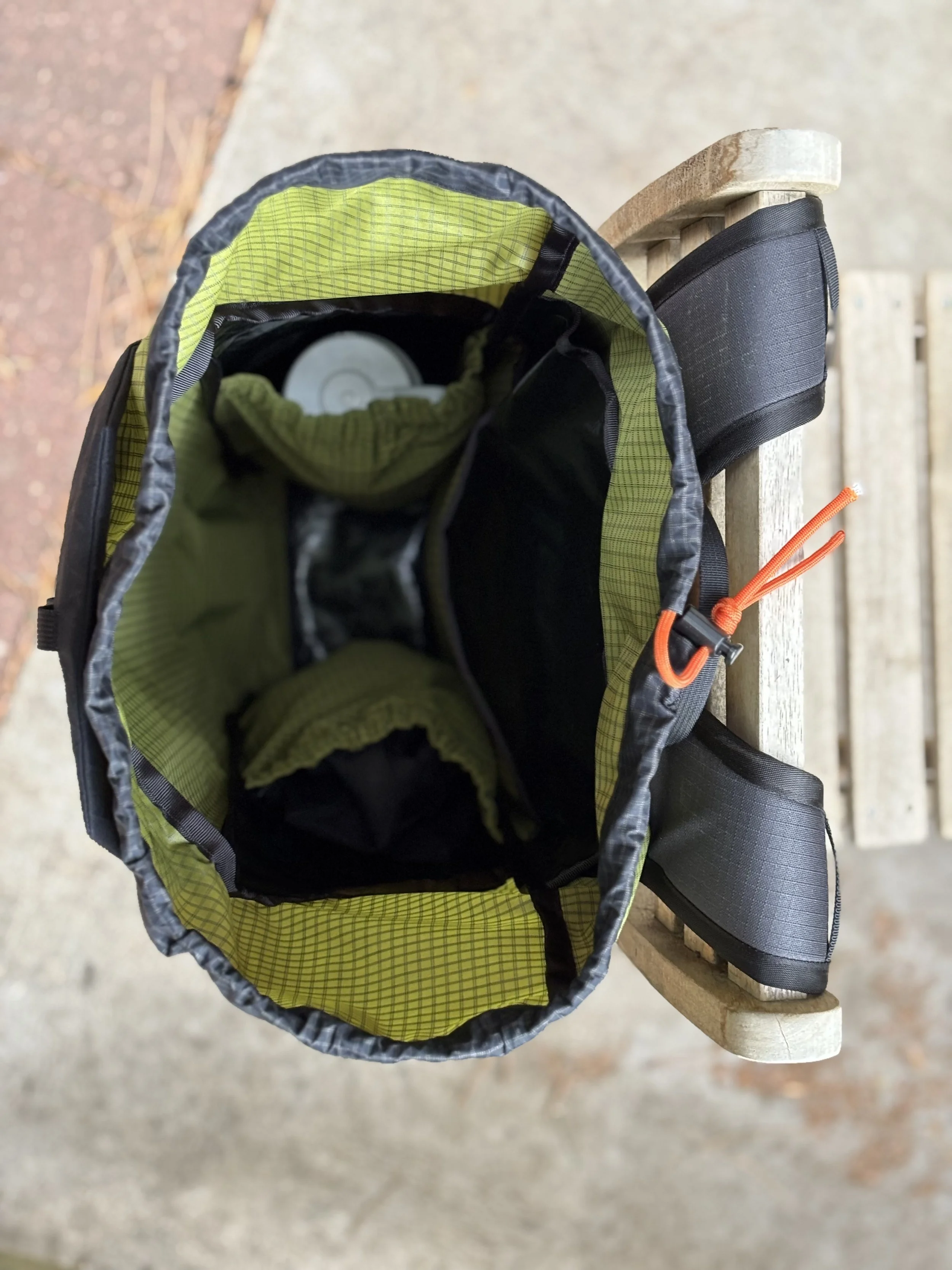 Open green-and-black backpack hanging on a wooden railing, containing a water bottle and clothing