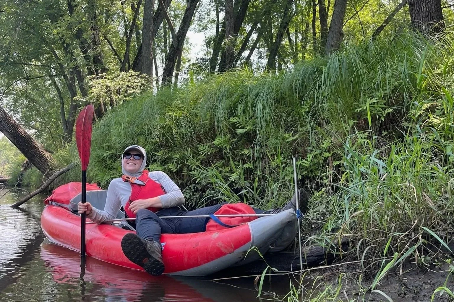 Smiling kayaker paddling a red inflatable kayak along a tree-lined waterway.