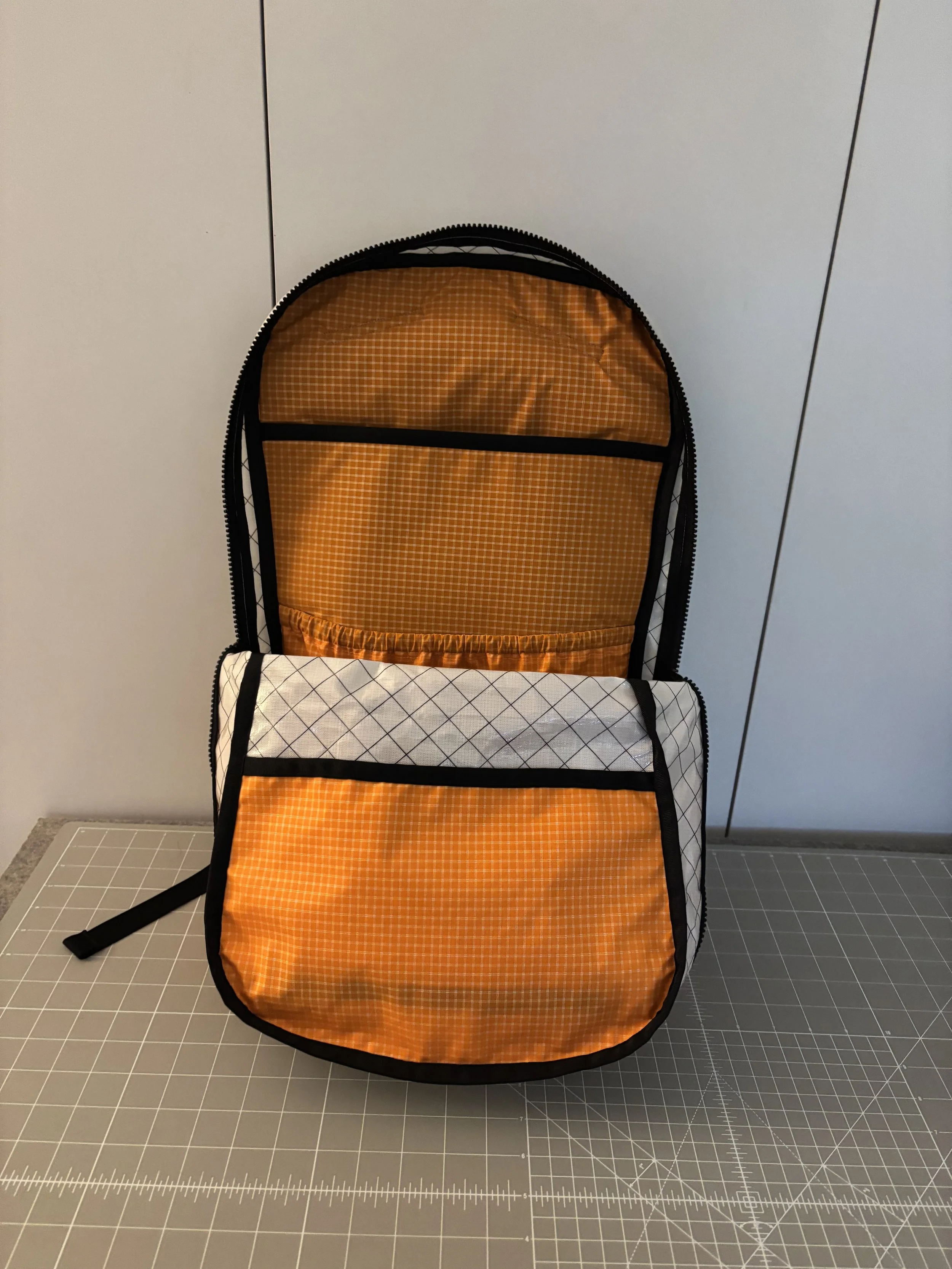 Orange plaid fabric backpack with black trim and white diamond-patterned side pockets on a cutting mat