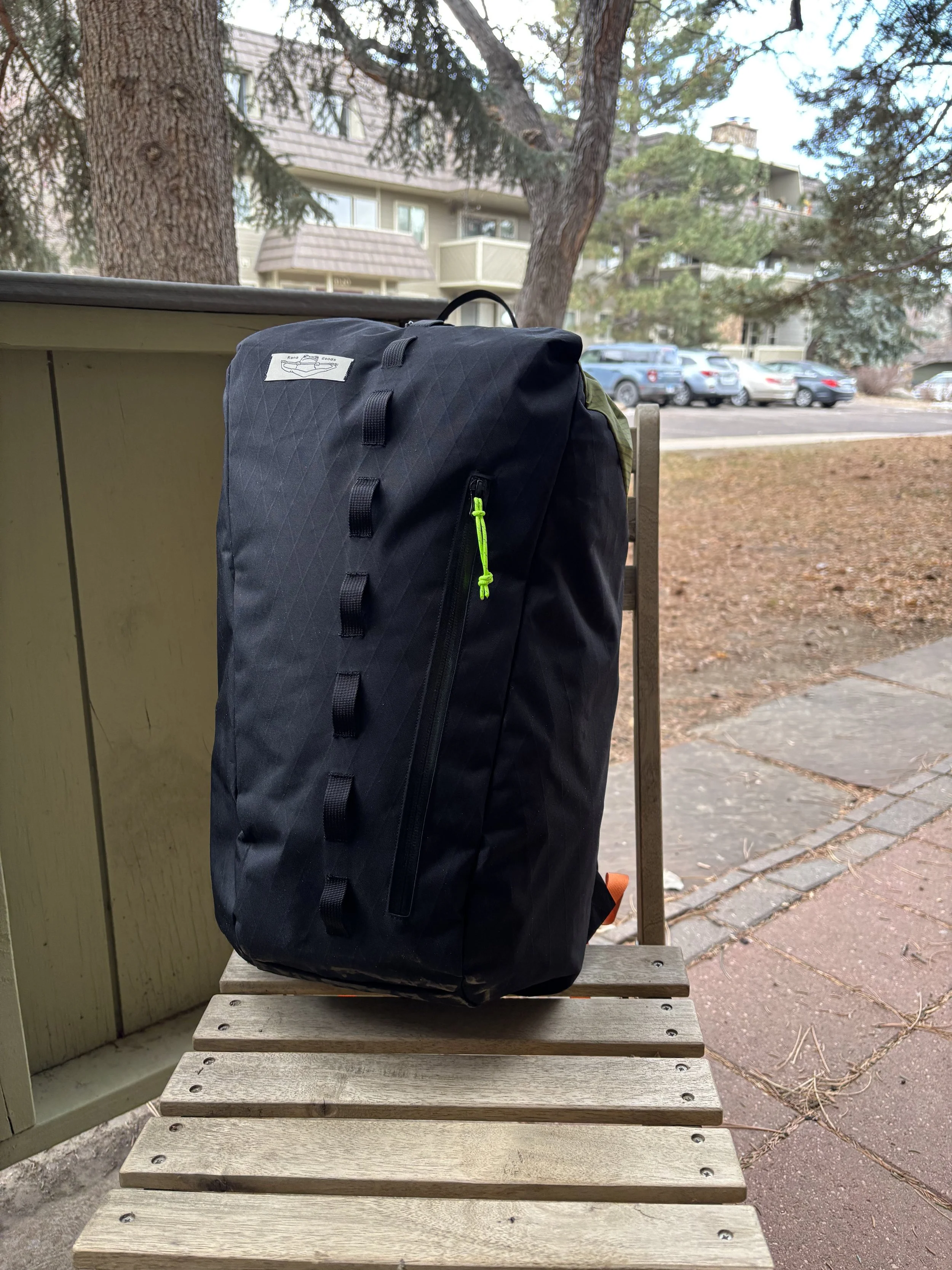Black backpack sitting on wooden outdoor steps beneath trees, with apartment buildings and parked cars in the background