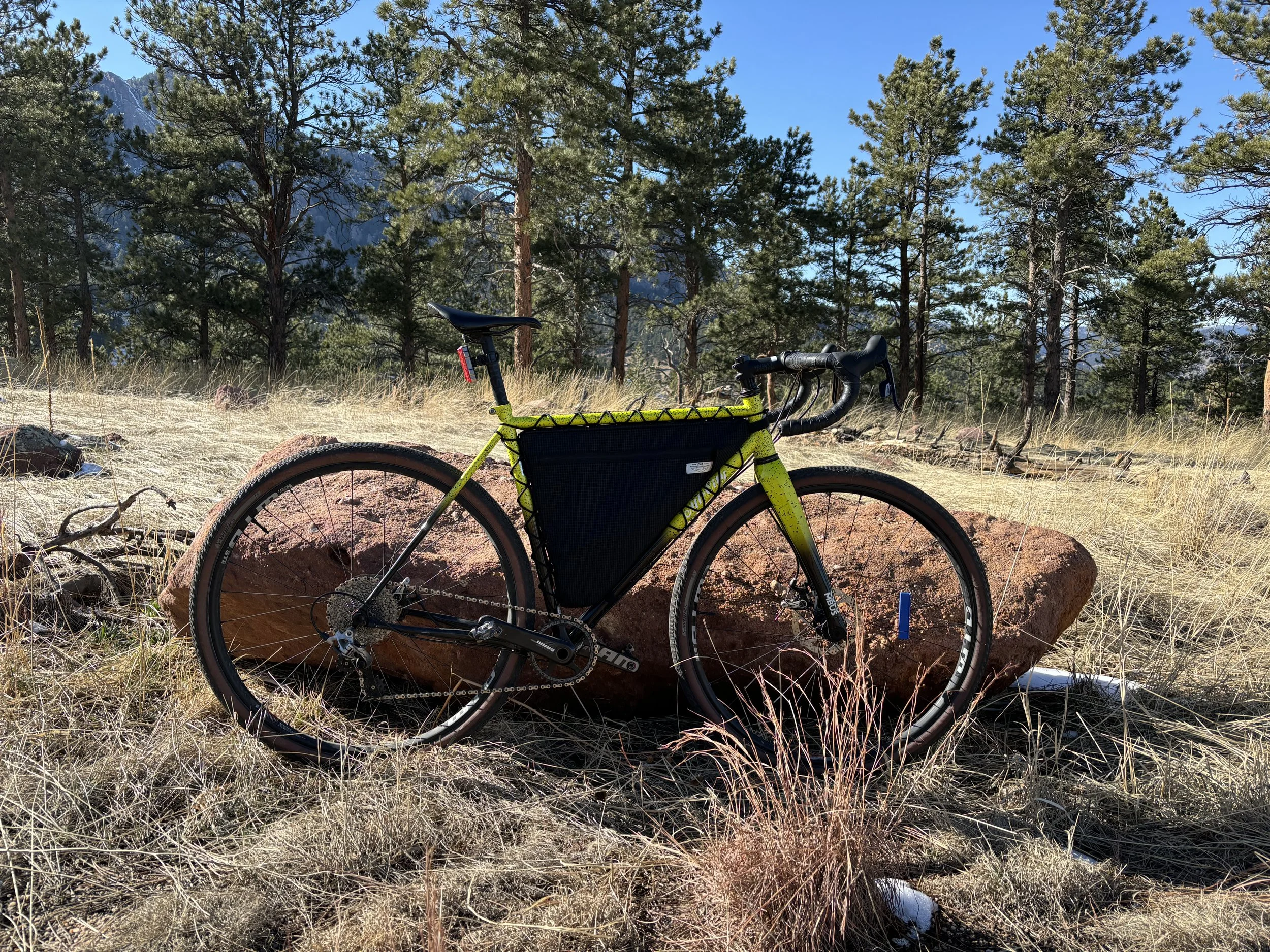 Yellow gravel bike with a black frame bag parked among pine trees and boulders, with Rocky Mountain peaks in the background.
