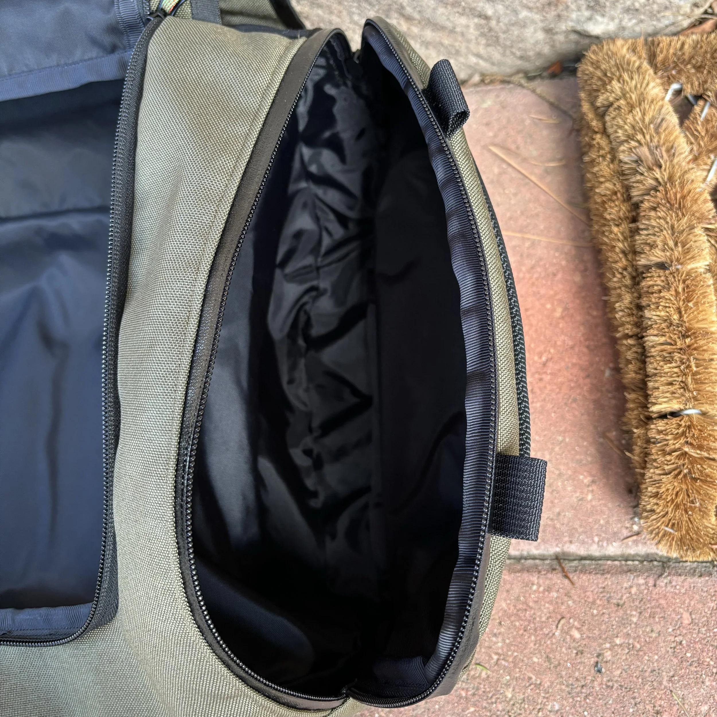 Open olive-green backpack with a black interior beside a brown furry dog on a brick patio