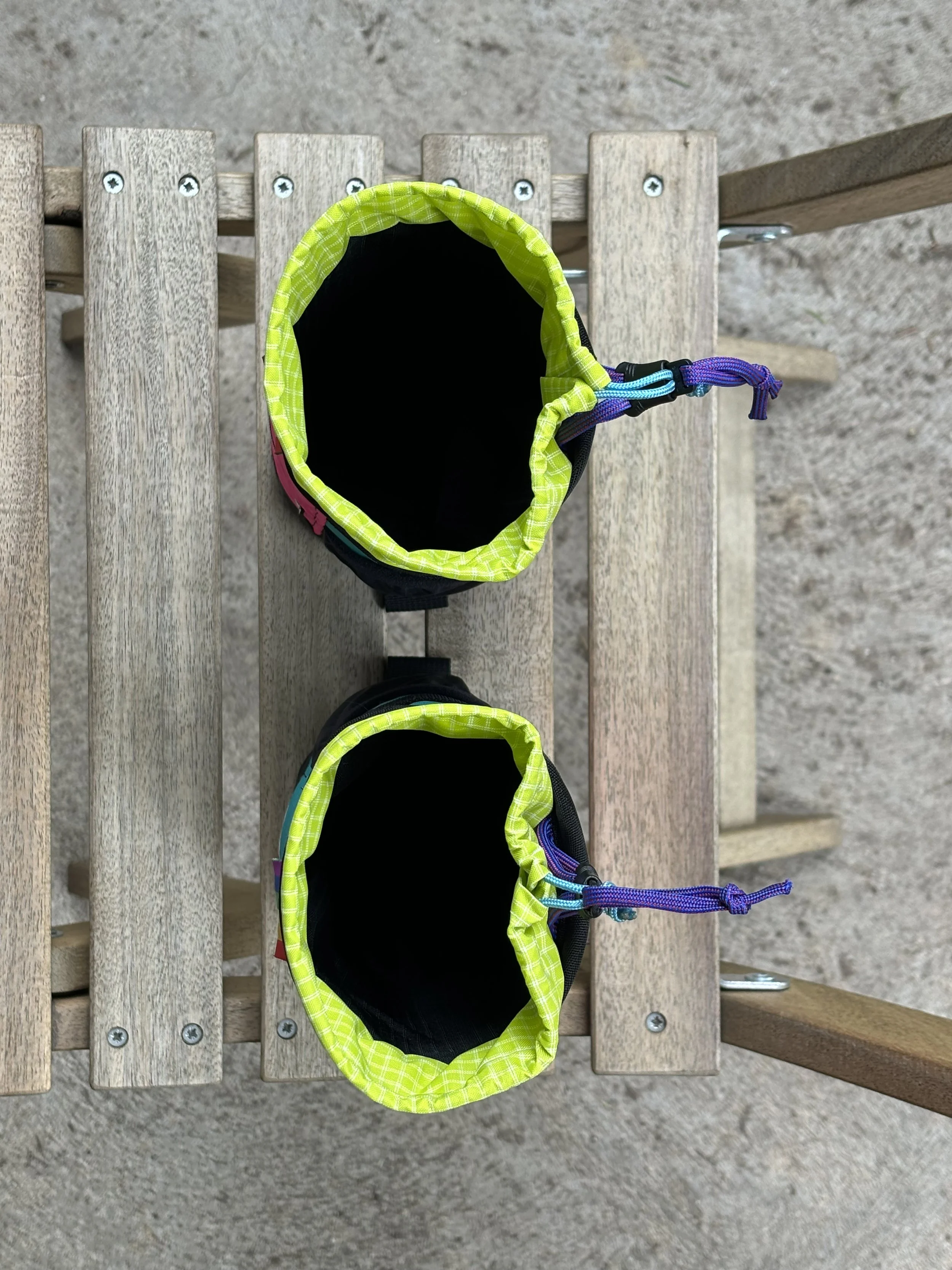 Two neon green-trimmed black pet backpacks with purple and blue straps on a wooden bench