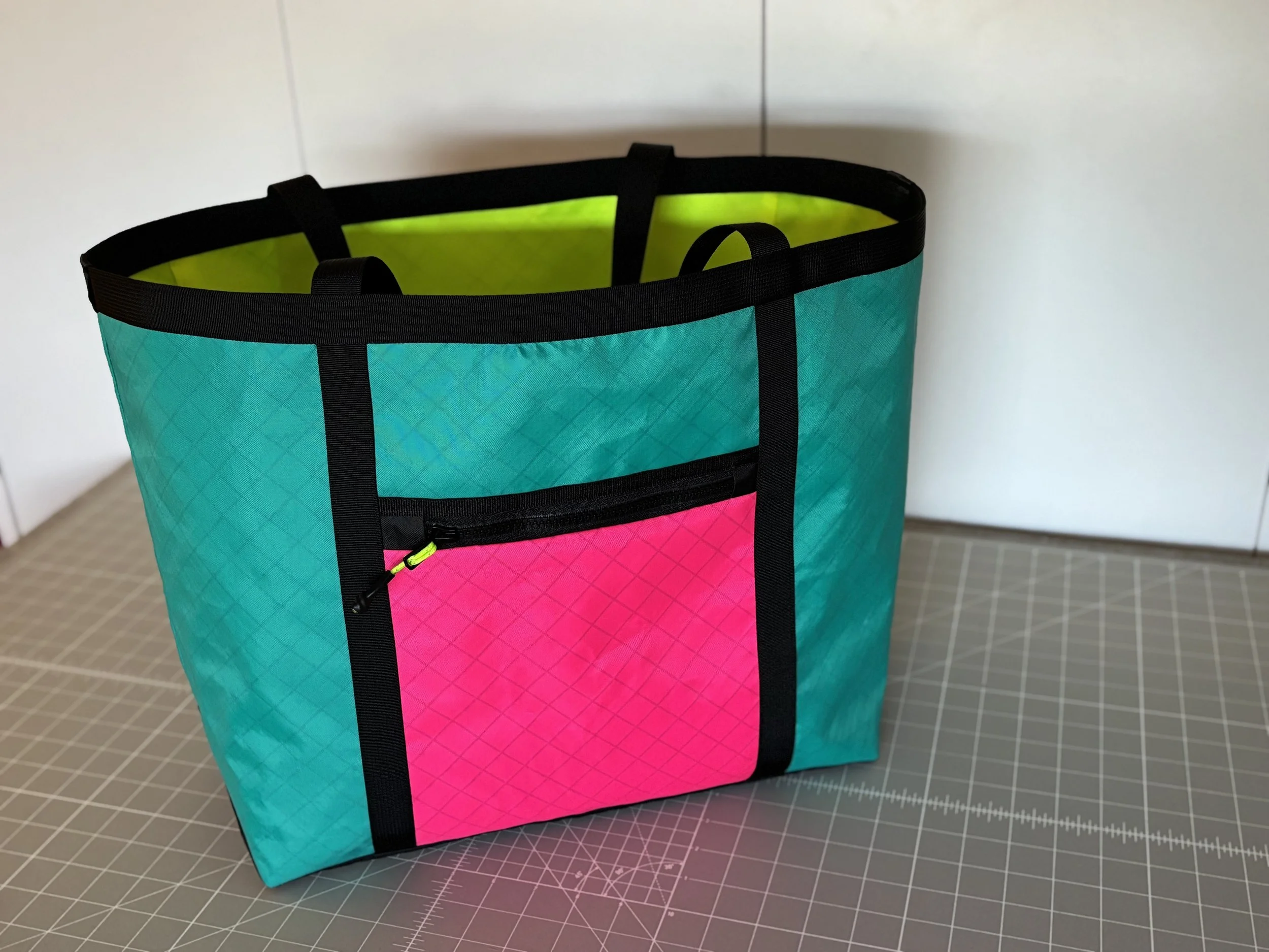 Colorful turquoise tote bag with black straps, neon pink zippered front pocket, and lime green interior