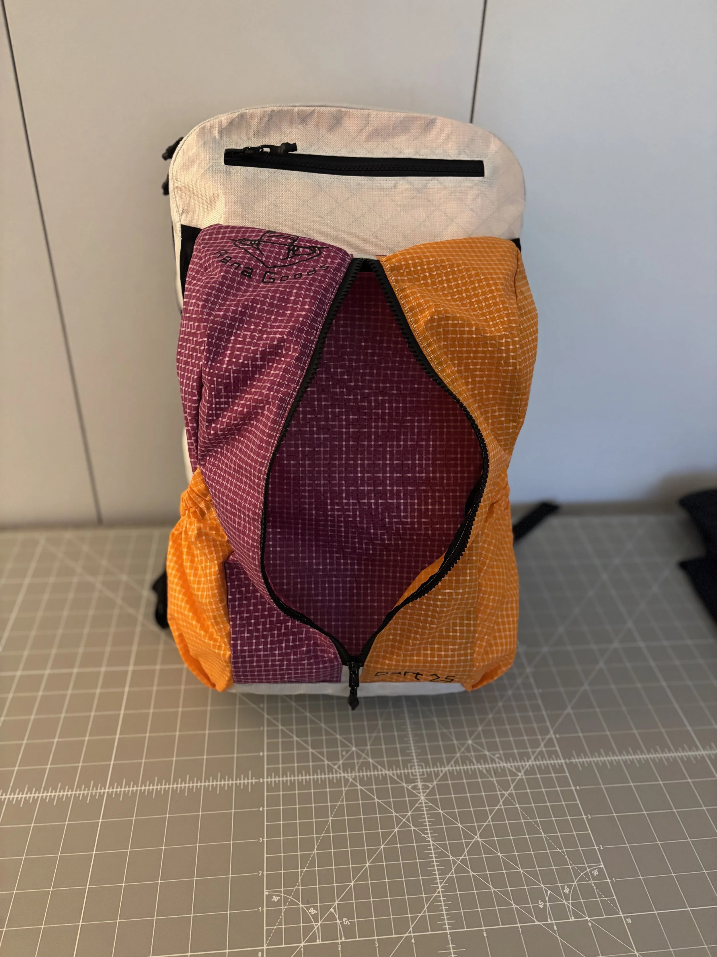 Colorful handmade patchwork backpack with orange and burgundy plaid panels, black trim, and a front zippered pocket on a cutting mat