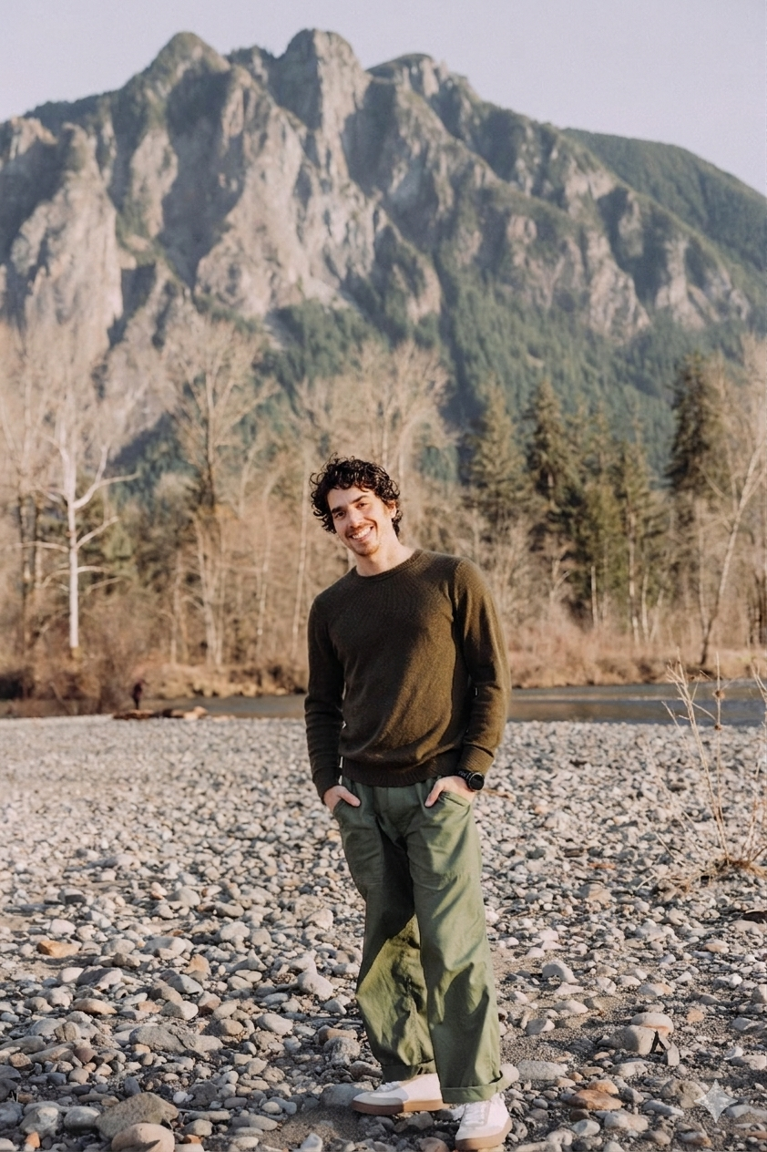 Smiling man standing on a rocky riverbank with a rugged, forested mountain behind him
