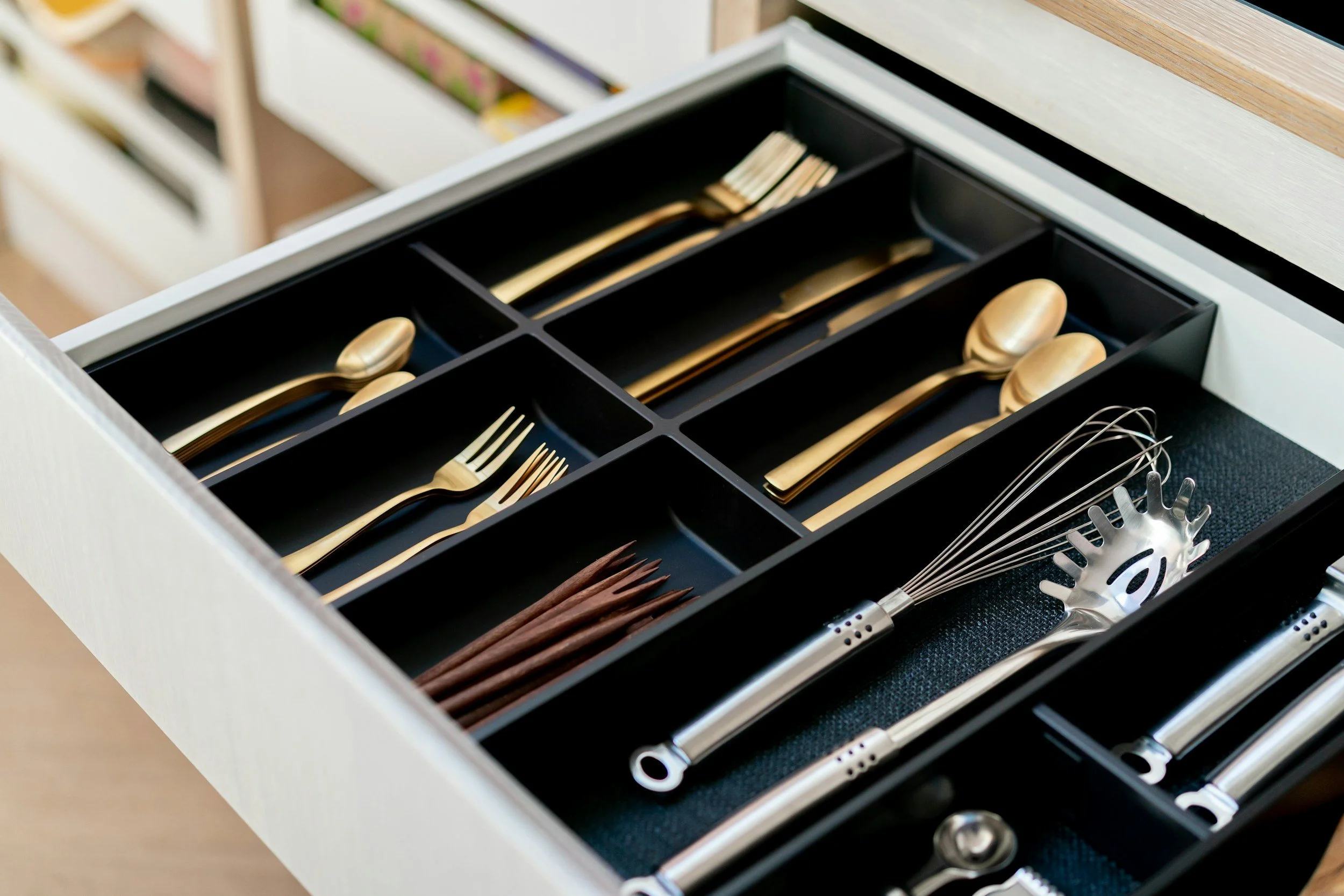 Open kitchen drawer with organized gold flatware, wooden chopsticks, and stainless steel utensils in black dividers