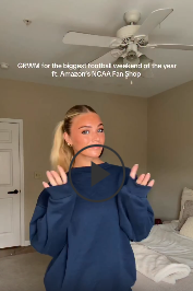 Smiling blonde woman in a navy sweatshirt holding a hanger in a bedroom, with overlaid text promoting the NCAA Fan Shop.