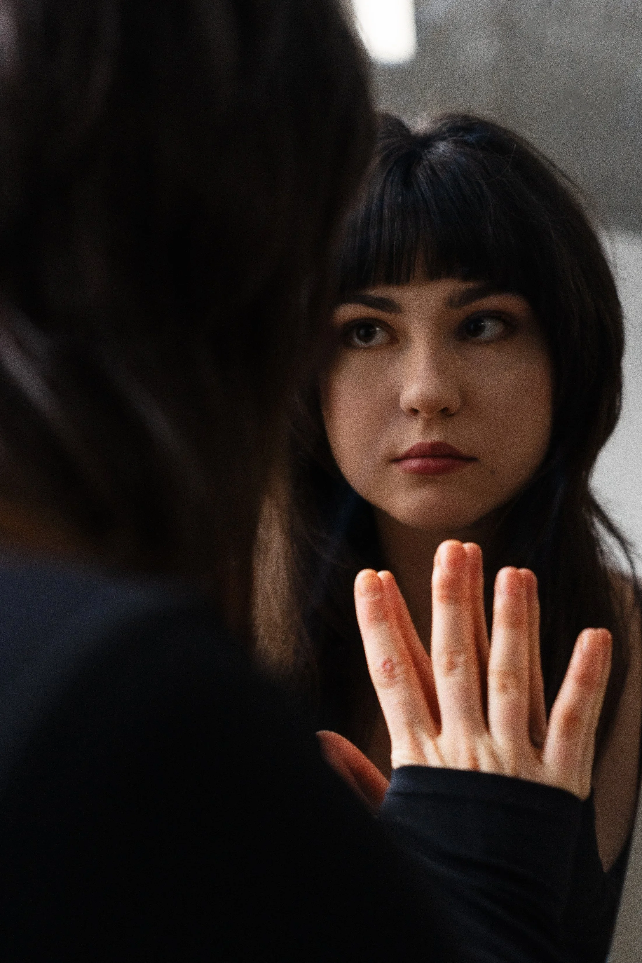 Close-up of a woman with dark hair looking into a mirror while touching her reflection