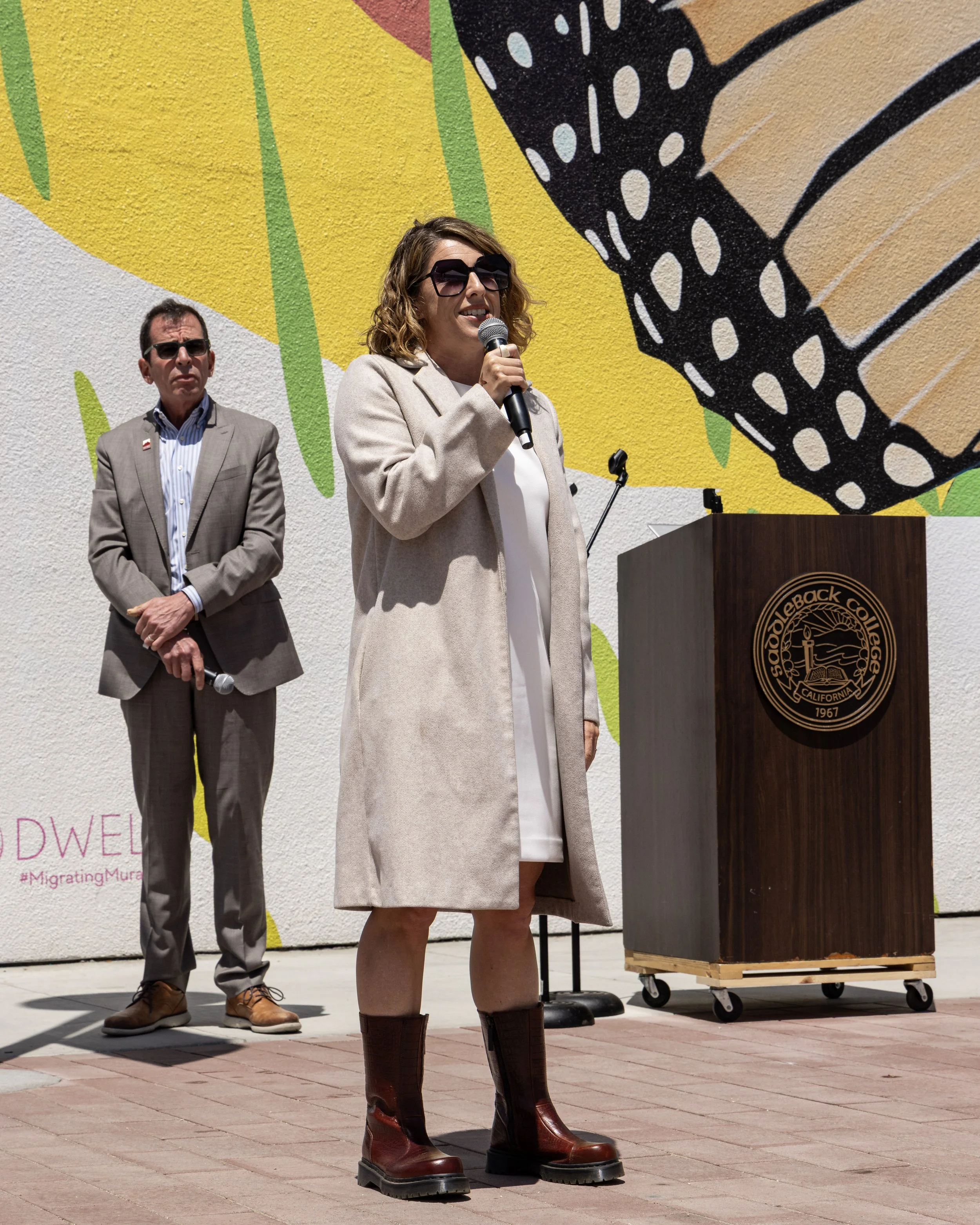 Woman speaking into a microphone beside a man and podium in front of a colorful mural.