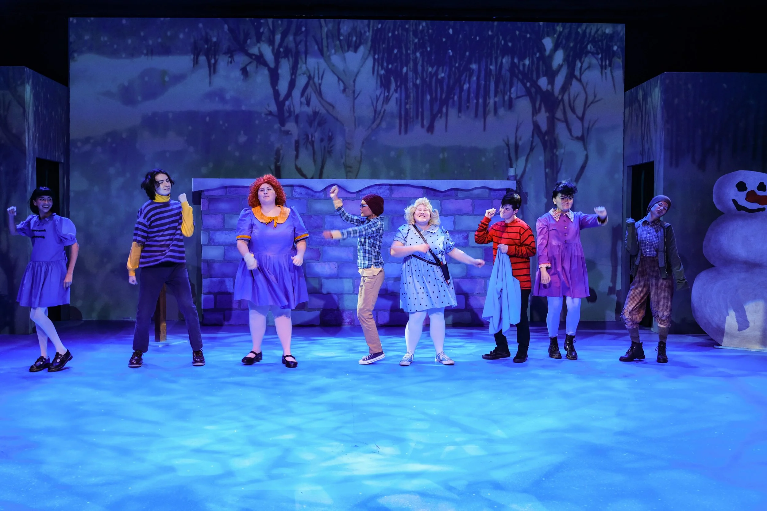 Cast members perform on a blue-lit stage in colorful costumes beside a stone wall and large snowman prop.