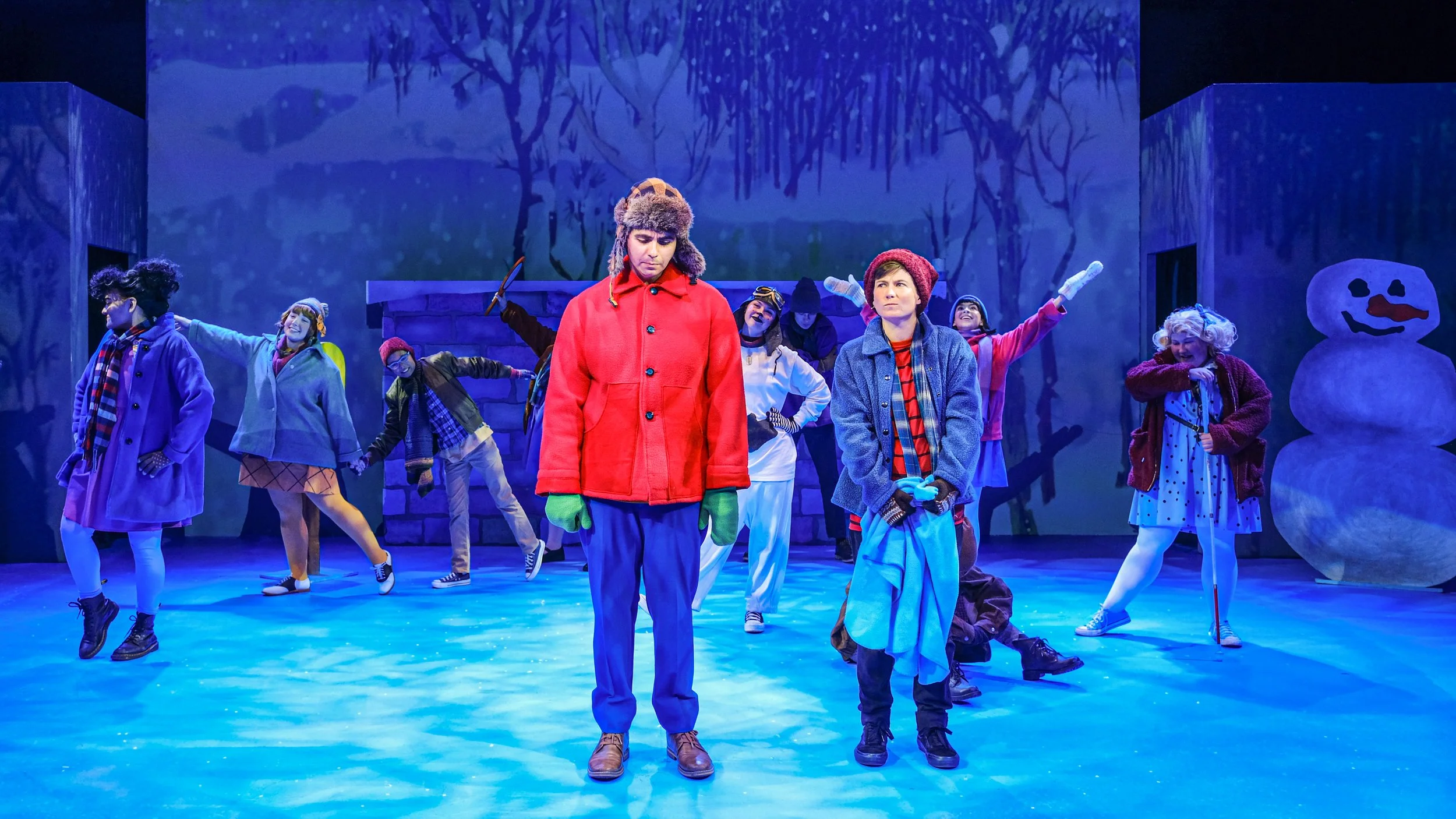 Cast members in colorful winter costumes perform on a blue-lit theater stage beside a large snowman prop.