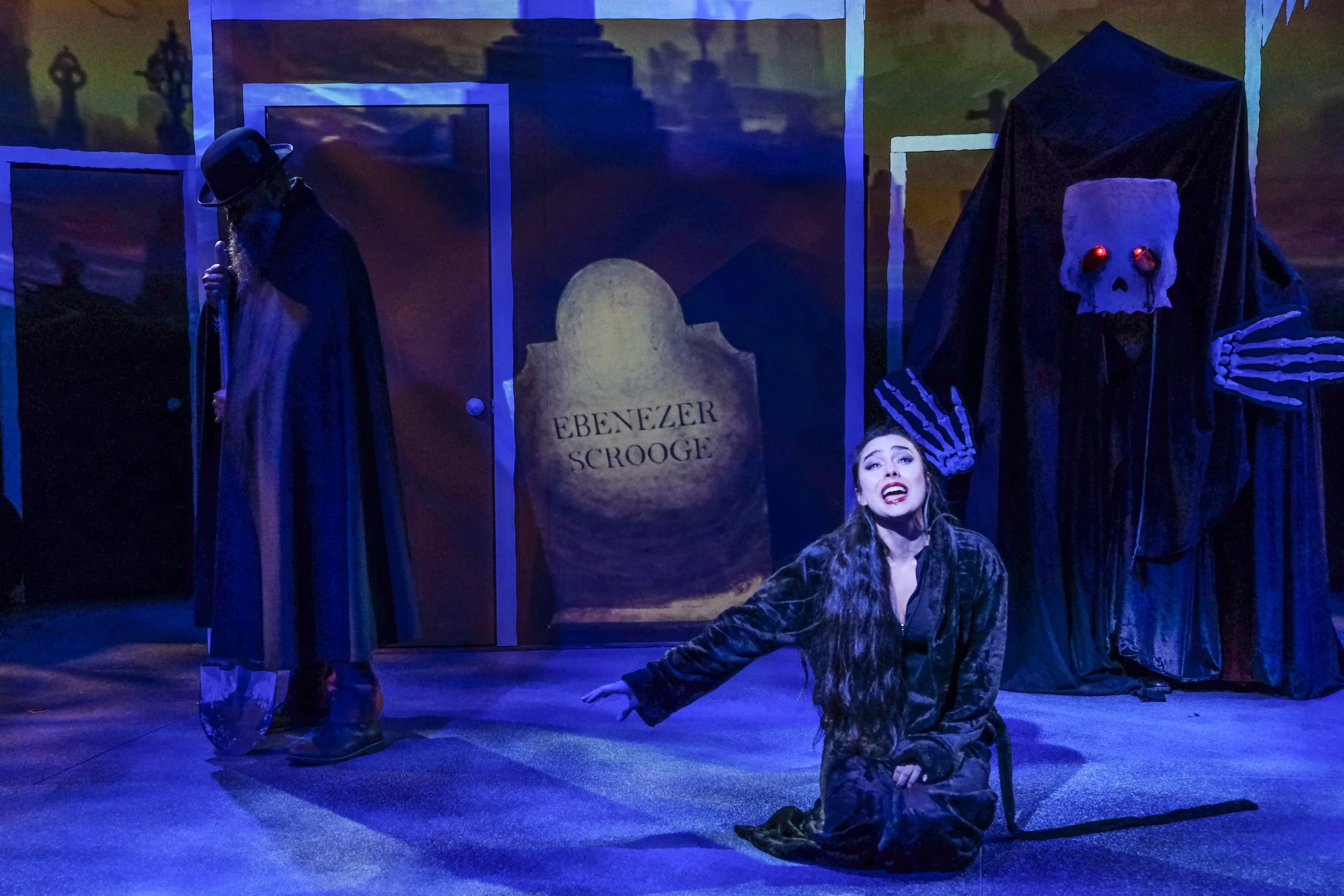 Theater scene featuring a woman in a black costume, a cloaked figure, and a glowing-eyed skeleton beside Ebenezer Scrooge’s tombstone.